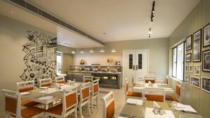Un comedor con mesas blancas y sillas con cojines naranja, una cocina al fondo y cuadros en las paredes.
