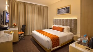 Habitación de hotel con cama, escritorio, computadora, silla, mesita de noche y un cuadro colgado en la pared.