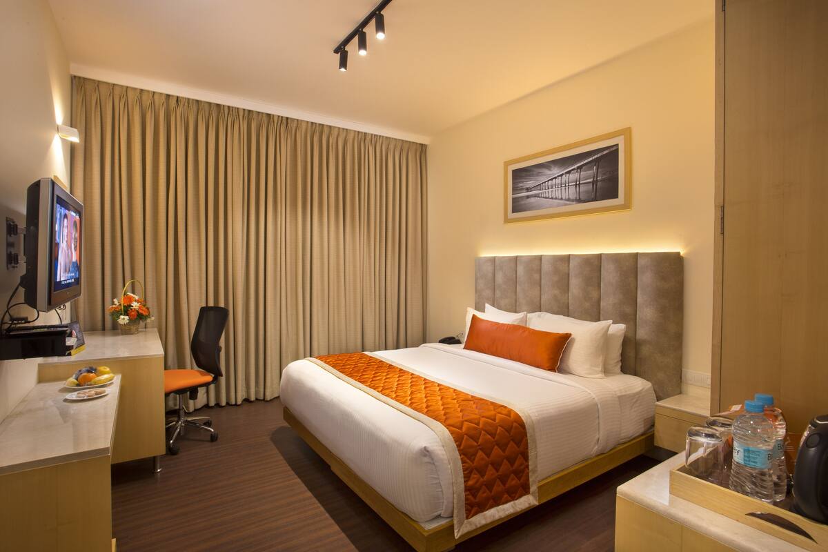 A hotel room with a bed, a desk with a computer, a chair, a nightstand, and a wall-mounted picture.