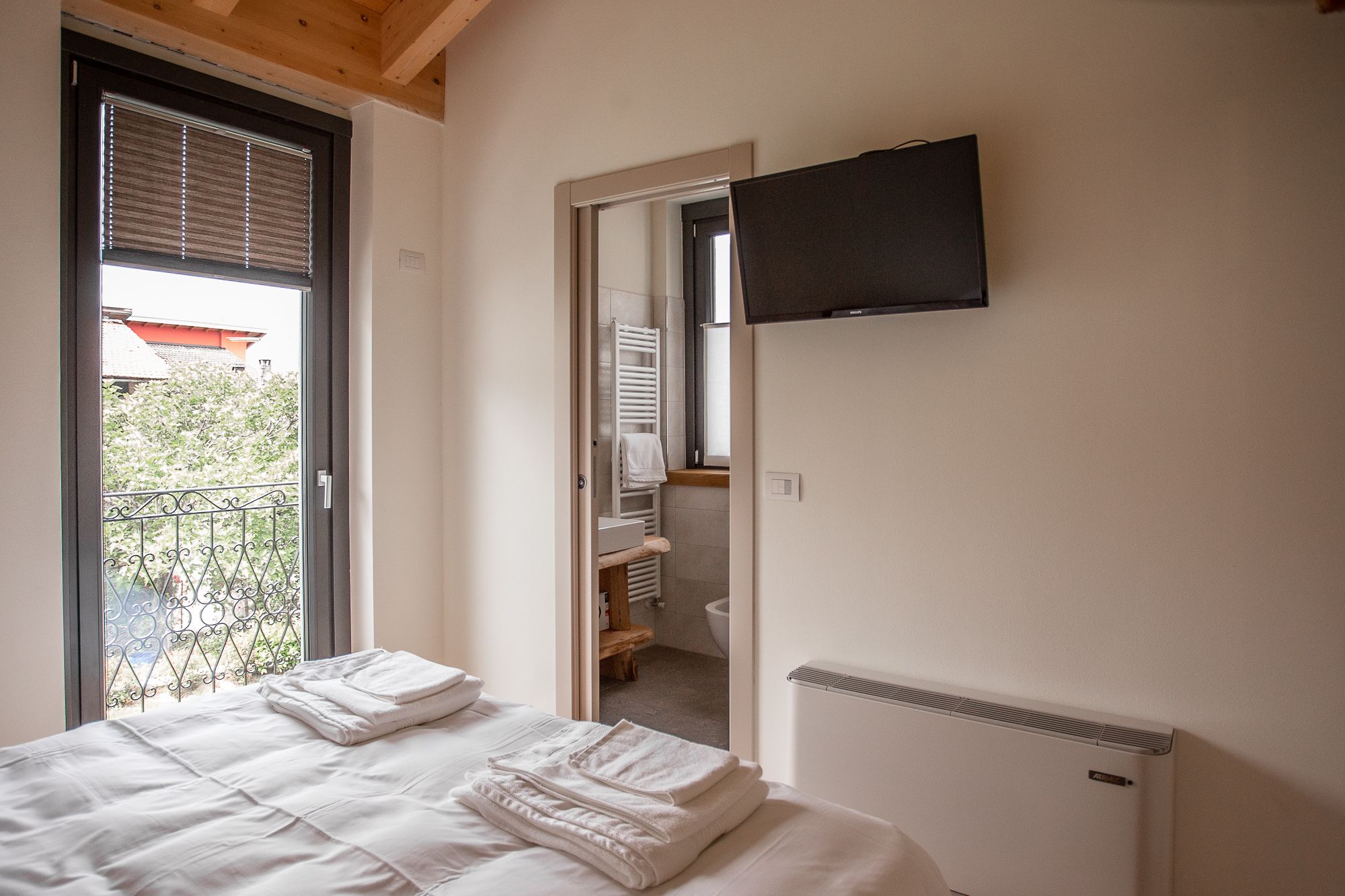 Un dormitorio con cama, radiador, televisor montado en la pared y vista al balcón exterior.
