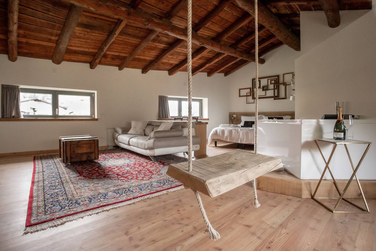 Ein geräumiges Wohnzimmer mit Holzbalkendecke, einer Schaukel, einem Sofa, einem Bett und einem kleinen Tisch mit Flasche und Gläsern.