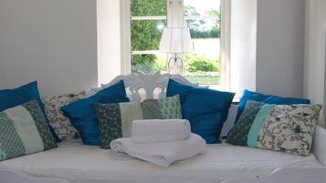 A bed with white bedding, multiple pillows in various patterns, and a lamp near a window.