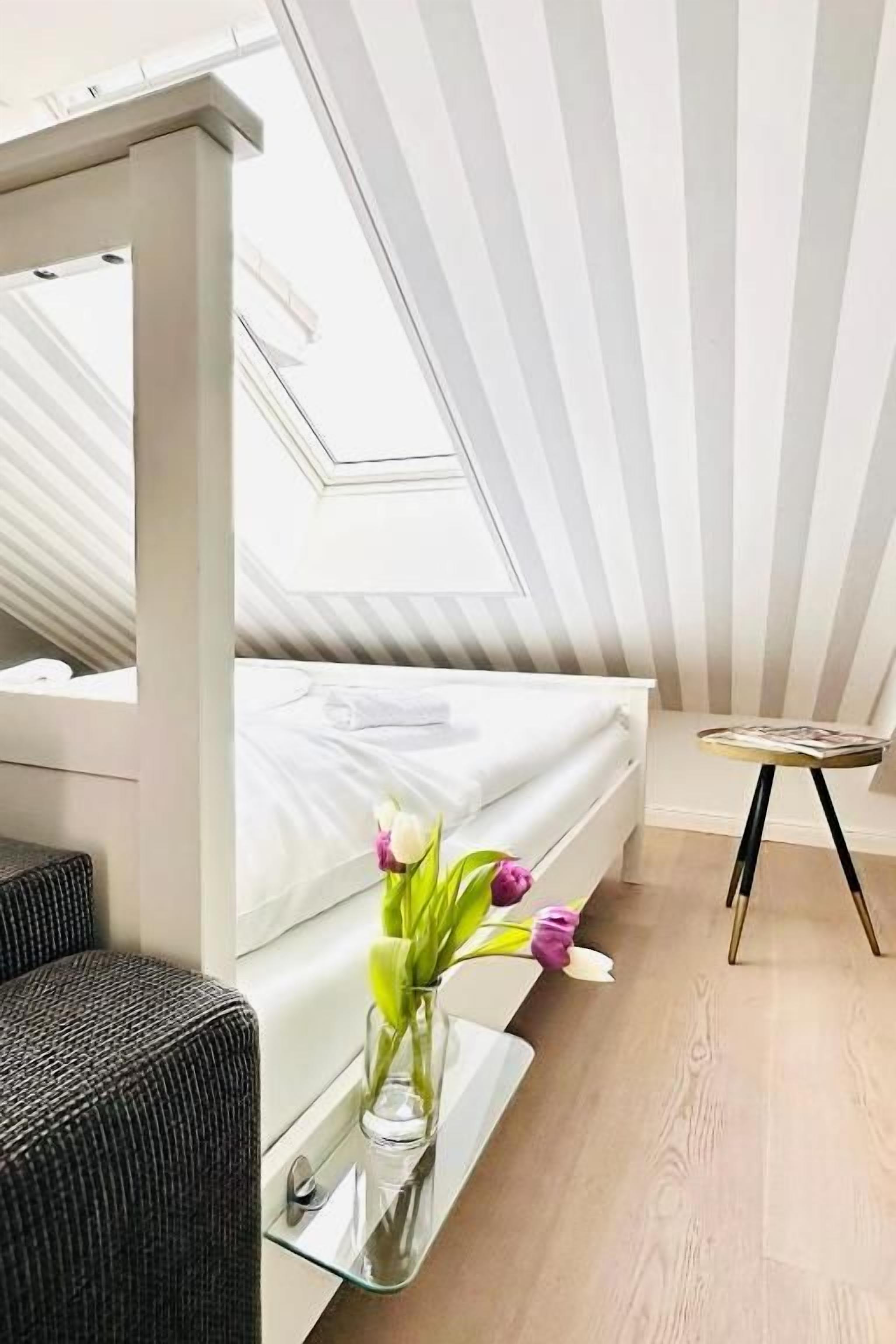 Ein Zimmer mit einem Bett, einem kleinen Tisch mit Blumenvase und einem Oberlicht.