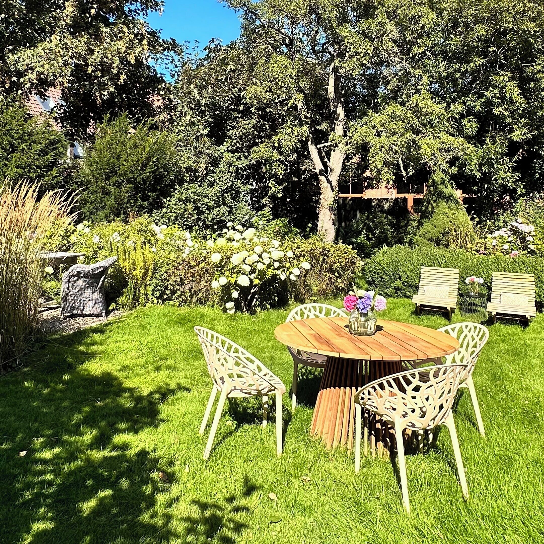 An outdoor seating area with a wooden table and white chairs, surrounded by lush greenery and flowering plants.