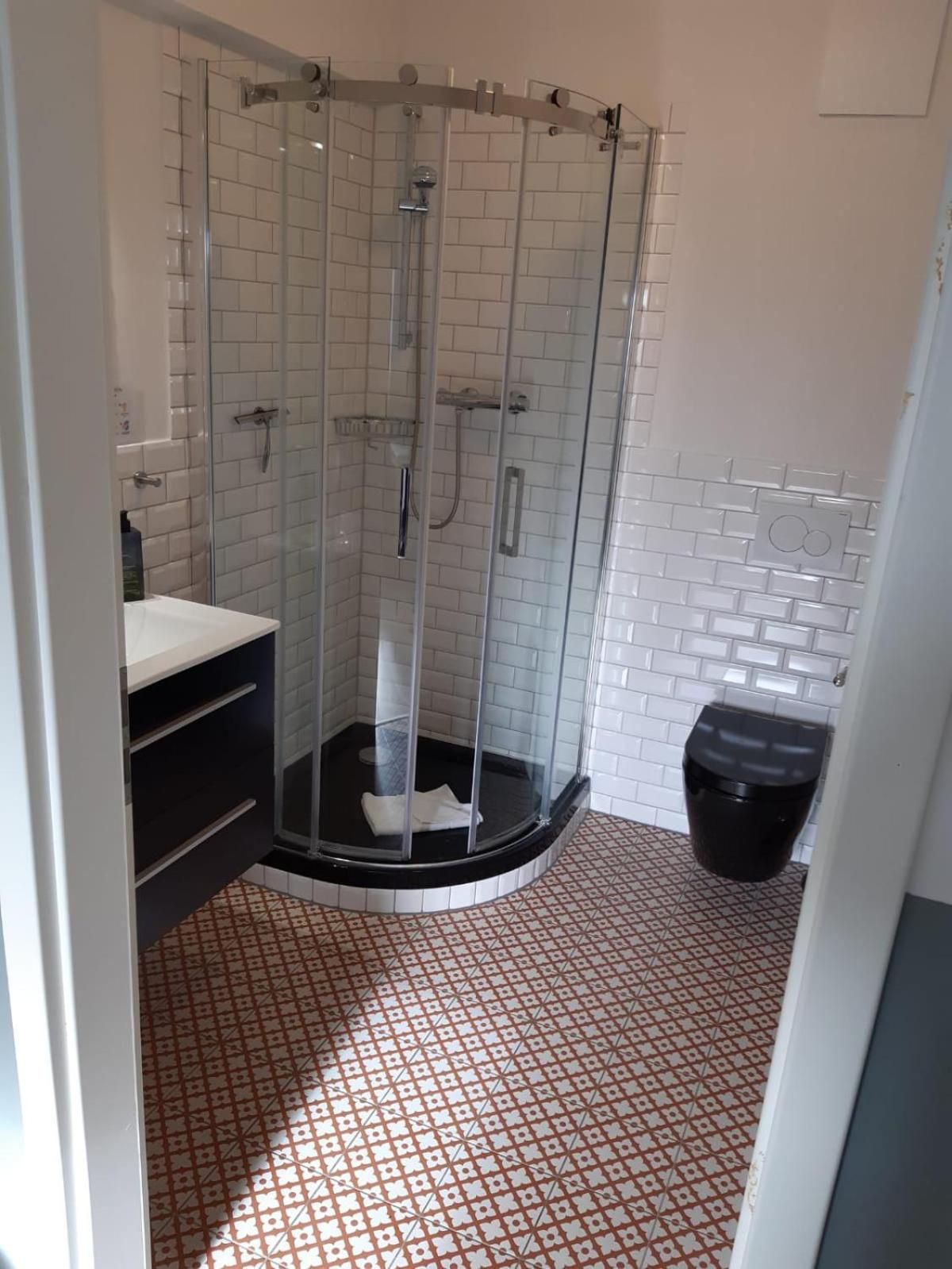 A shower with a glass enclosure, white tiled walls, a black trash bin, and patterned floor tiles.