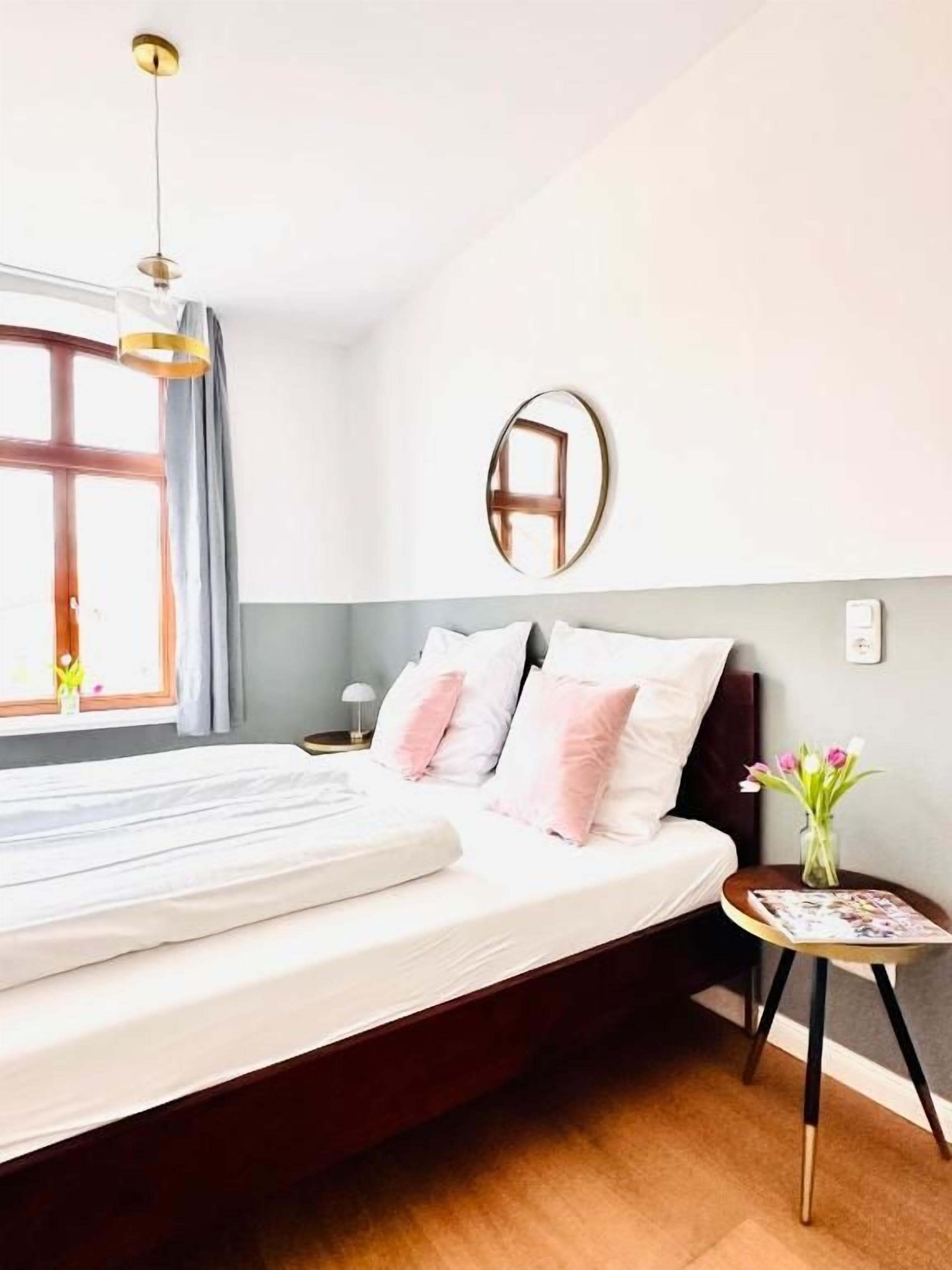 Un dormitorio con cama, mesita de noche, espejo y ventana con cortinas.