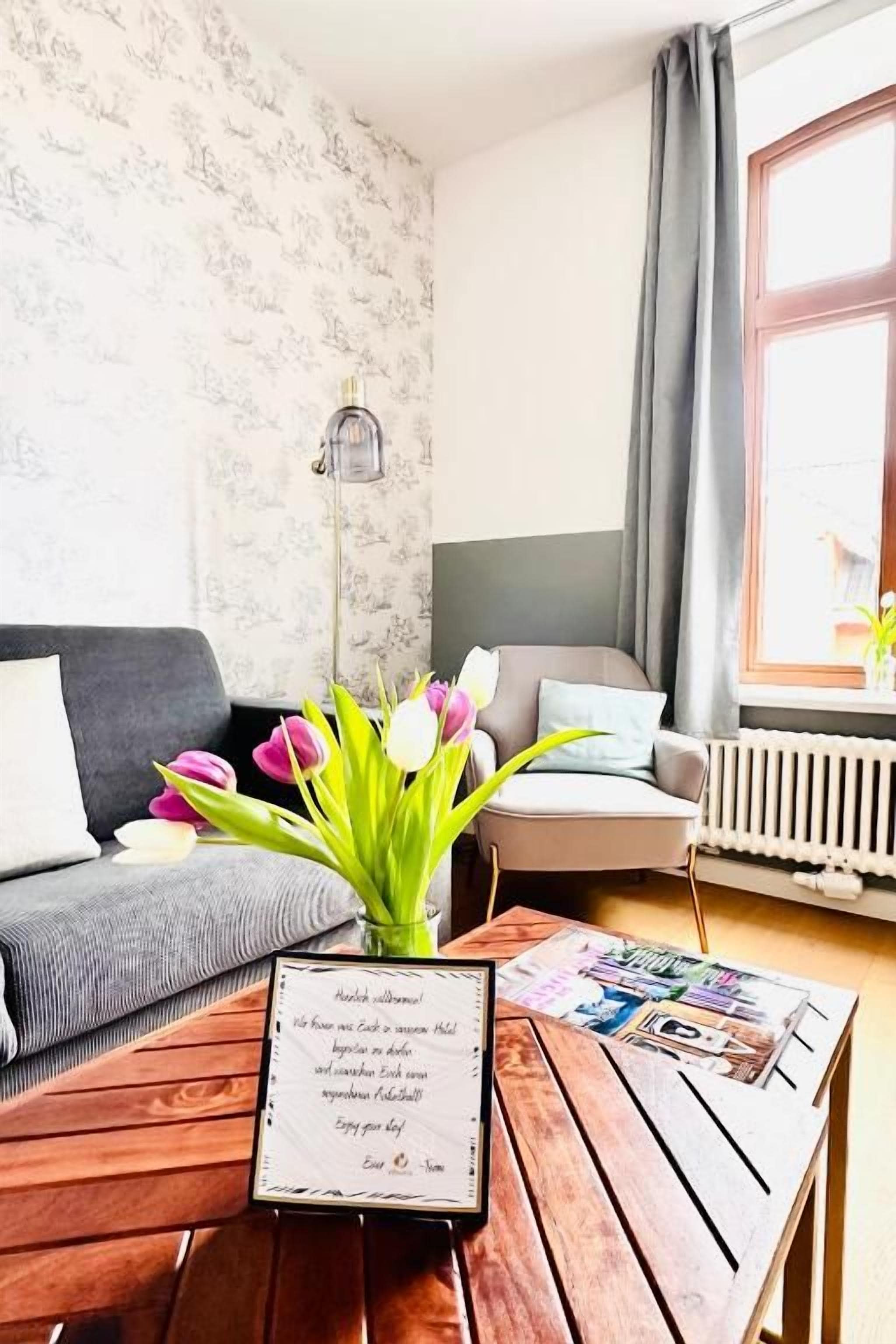 Een knus ingerichte kamer met een houten bijzettafeltje, een bord met daarop een welkomstboodschap, een grijze bank, een witte radiator en een raam met gordijnen.