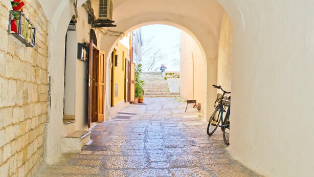 Um estreito beco de pedra com teto arqueado, ladeado por edifícios com portas e janelas de madeira. Uma bicicleta está estacionada à direita.