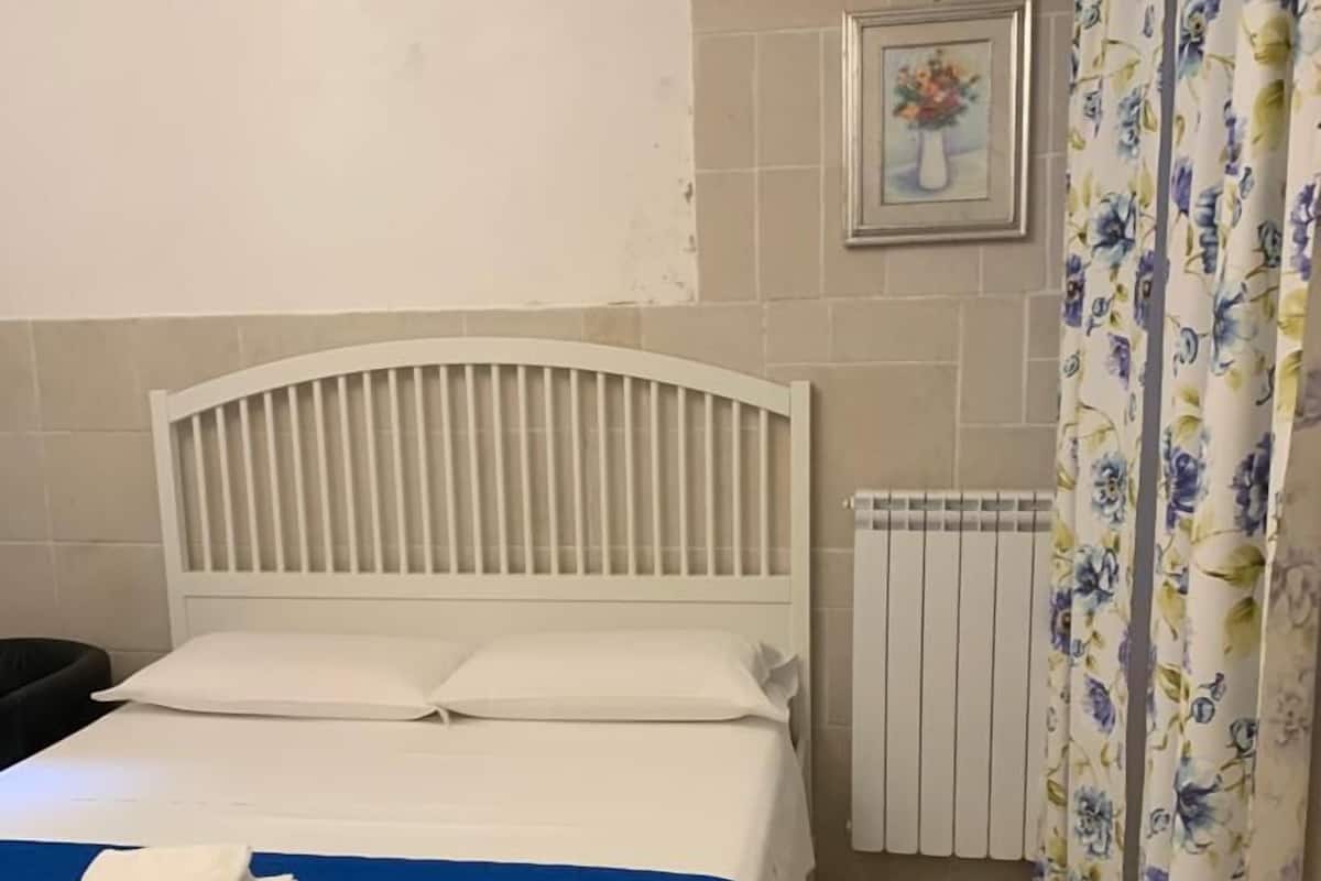 A neatly made bed with a blue bedspread and white pillows, a radiator, and a floral curtain.