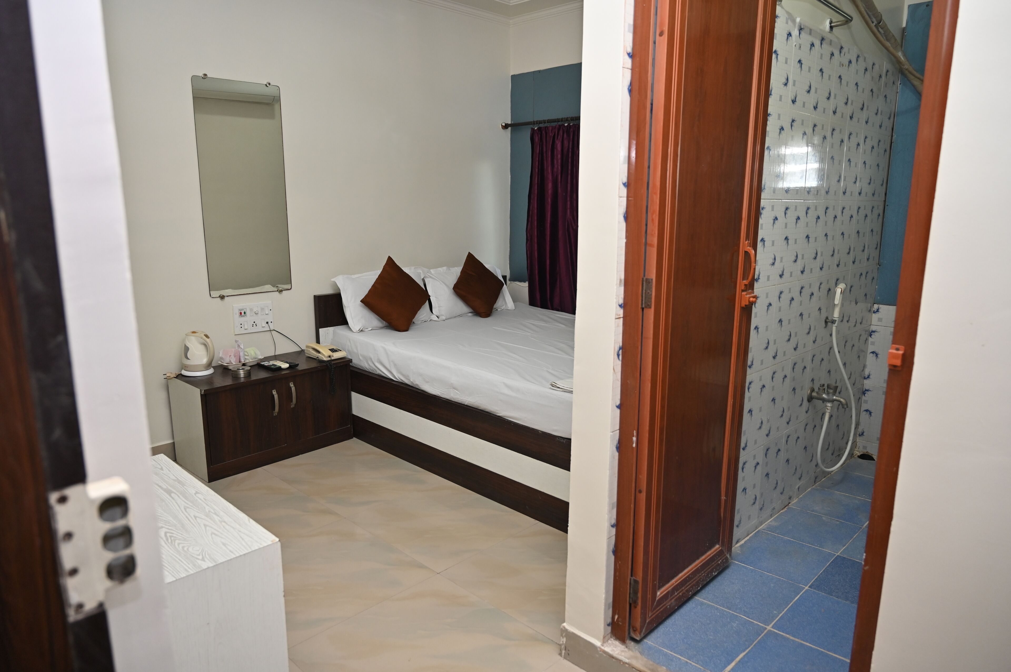 A bedroom with a bed, bedside table, and a mirror. There is a bathroom visible with a shower and a toilet.