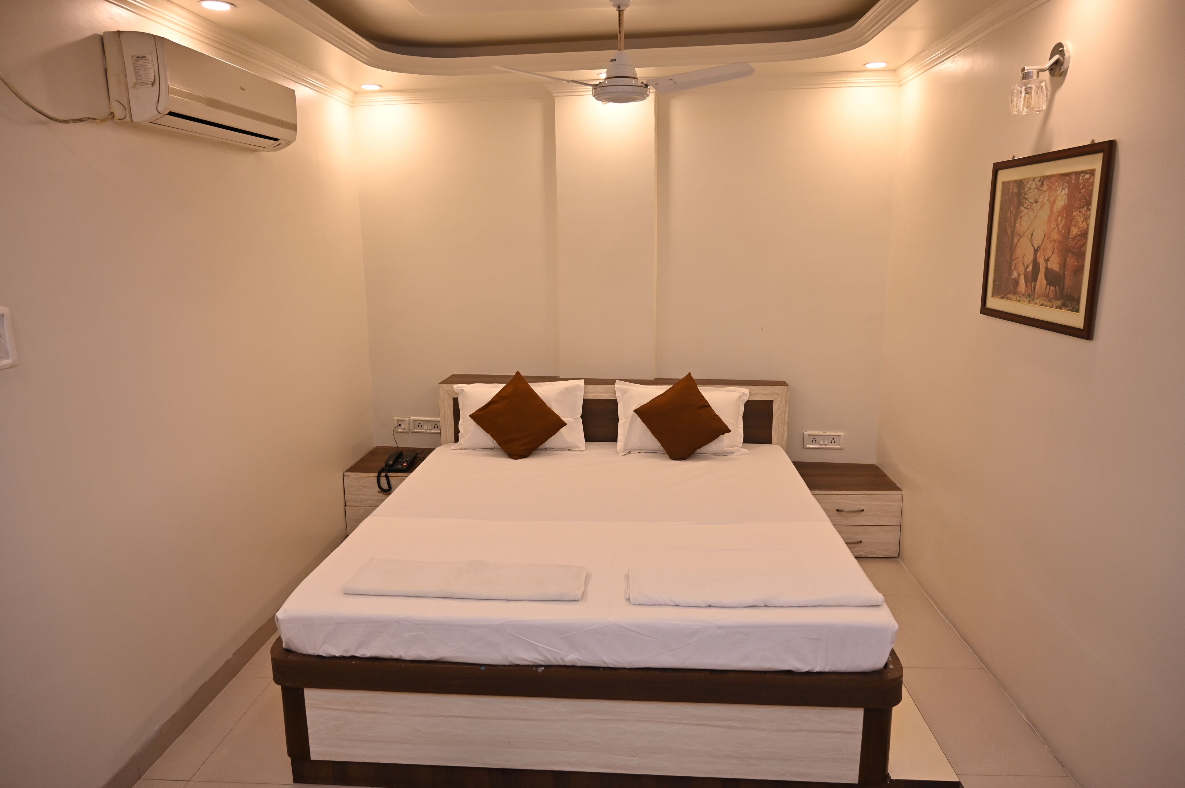 A neatly made bed with white linens and two brown pillows, a ceiling fan, and an air conditioning unit on the wall.