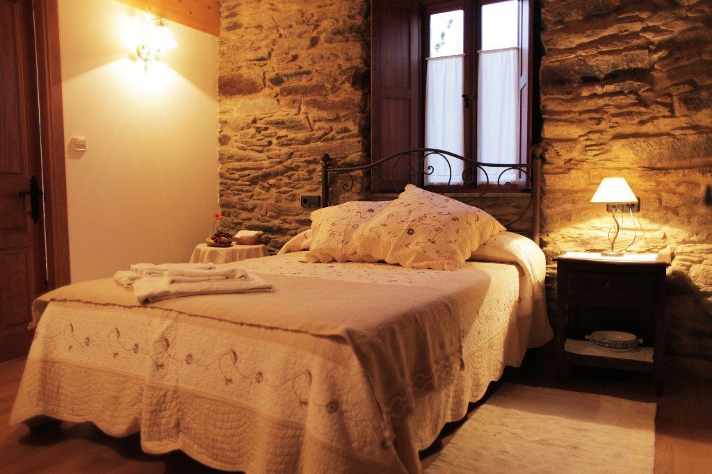 Un dormitori acollidor amb una paret de pedra, un llit amb estriat, una taula de nit amb una làmpada, i una finestra amb cortines.