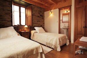 Ein gemütliches Zimmer mit zwei Betten, einer Steinwand, einer Holzbalkendecke, einem Nachttisch mit Lampe und einem Badezimmer mit Waschbecken und Spiegel.