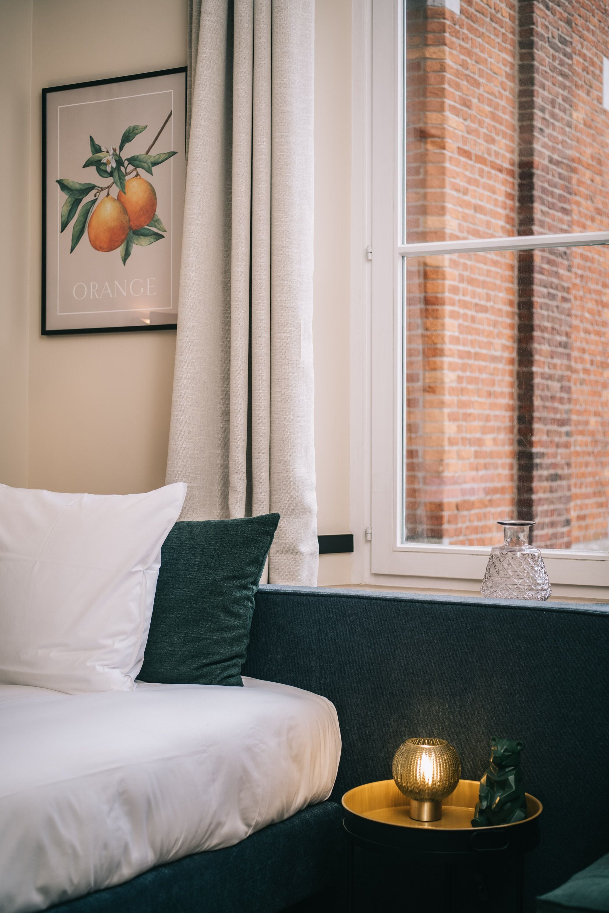Una cama bien hecha con almohadas blancas y verdes, un cuadro de fruta naranja enmarcado en la pared, una ventana con vista a un edificio de ladrillo y una mesita con una lámpara decorativa y una estatua de rana.