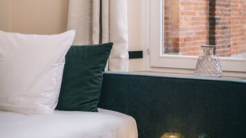 Una cama bien hecha con almohadas blancas y verdes, un cuadro de fruta naranja enmarcado en la pared, una ventana con vista a un edificio de ladrillo y una mesita con una lámpara decorativa y una estatua de rana.