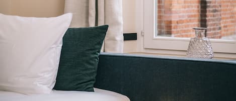 Een net opgemaakt bed met witte en groene kussens, een lijst met fruit in oranje tinten aan de muur, een raam met uitzicht op een bakstenen gebouw en een klein tafeltje met een decoratieve lamp en een kikkerbeeldje.