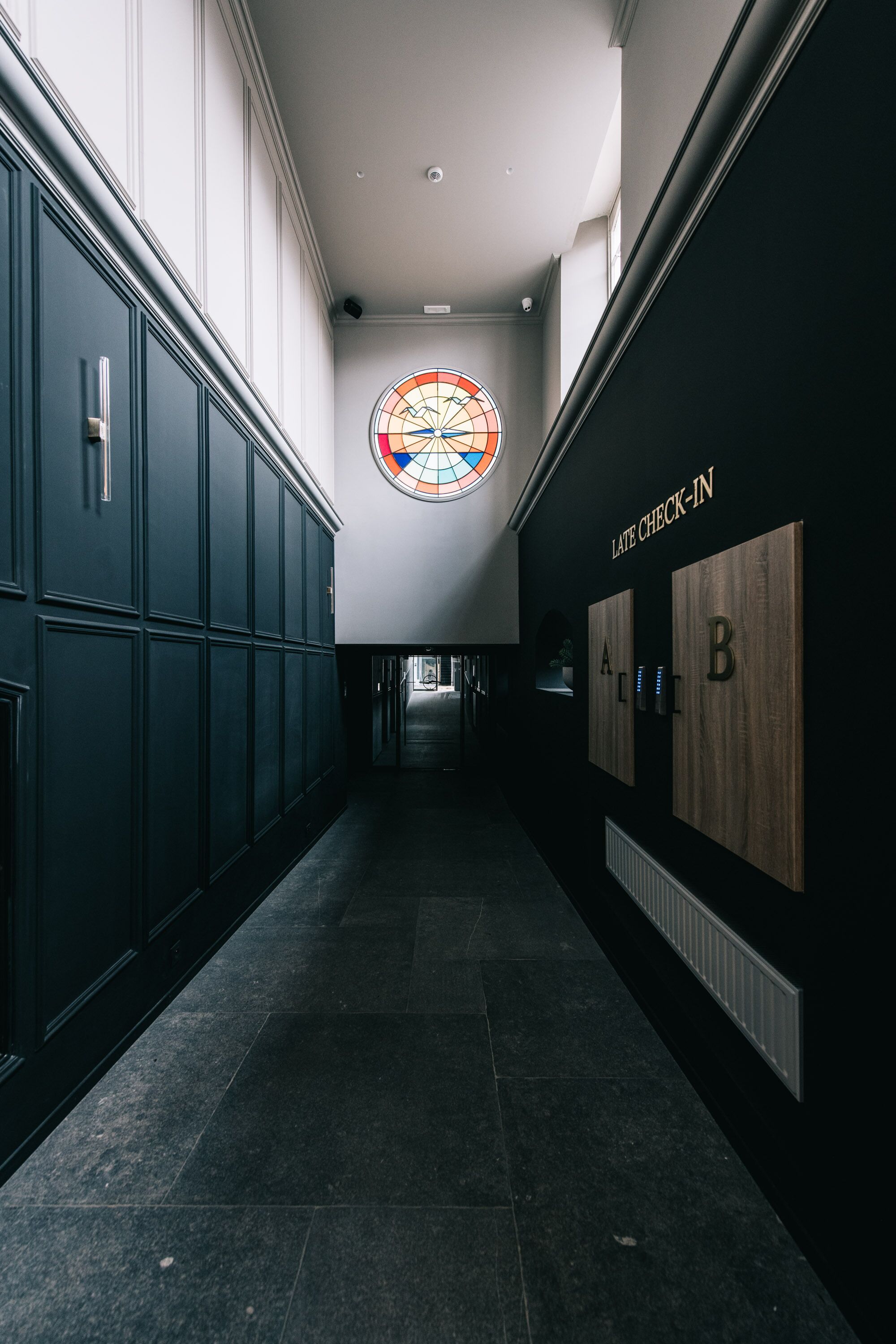 A hallway with lockers, a stained glass window, and signs for late check-in and room types A and B.