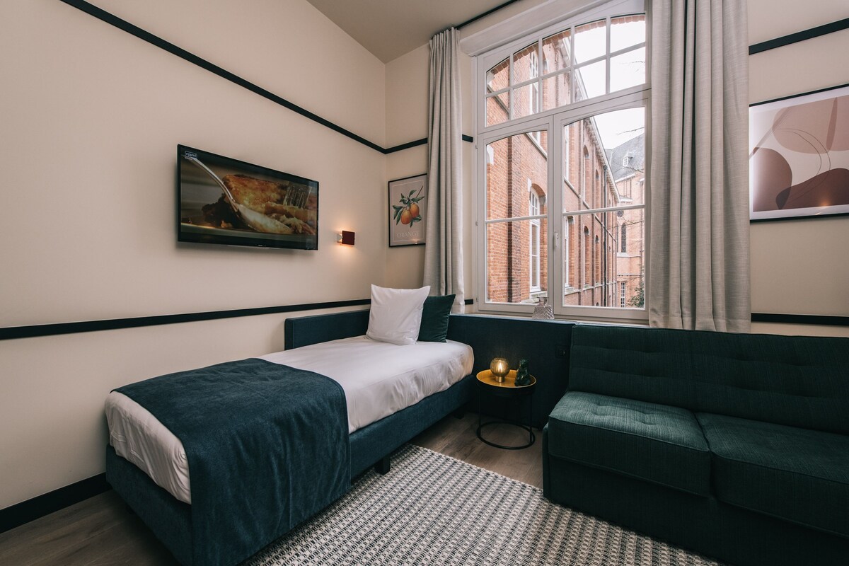 Un dormitorio con cama, sofá verde, ventana con cortinas y obras de arte en las paredes.