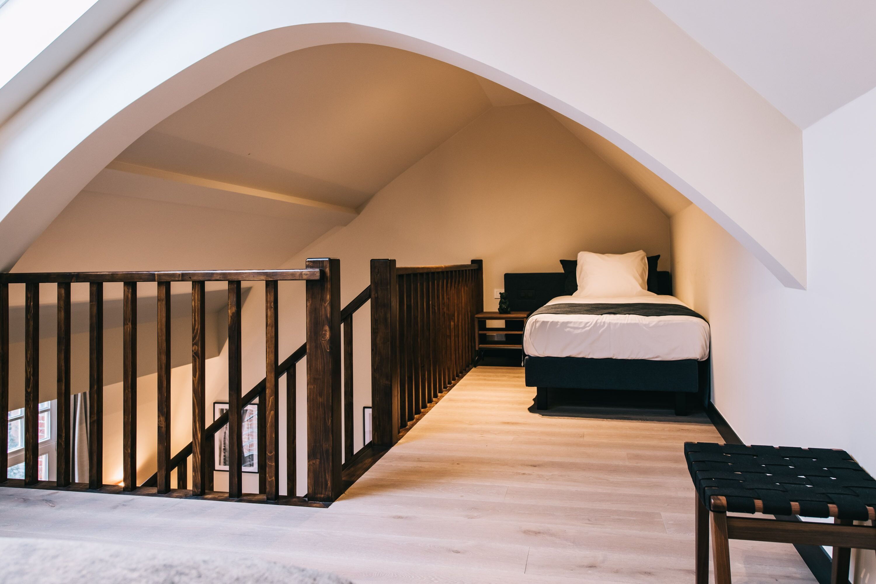 A bedroom with a wooden floor, a bed, a bench, and a staircase leading to an upper level.
