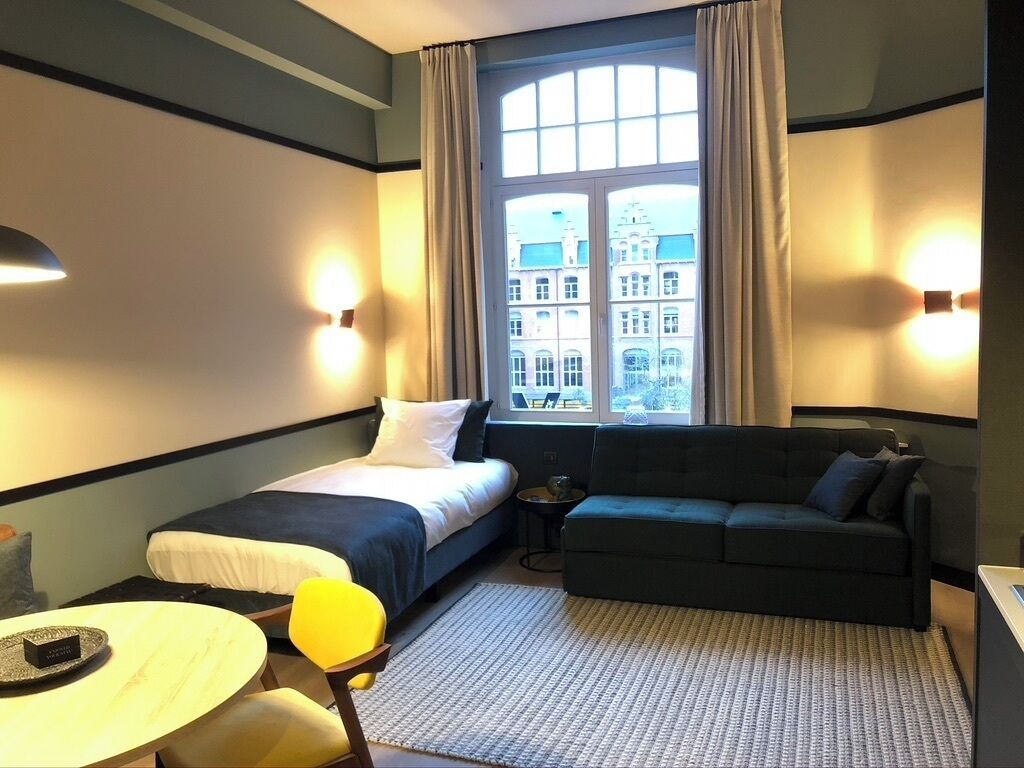 Ein Hotelzimmer mit Bett, einer Sitzecke, einem kleinen Tisch und Blick auf ein Gebäude durch ein Fenster.