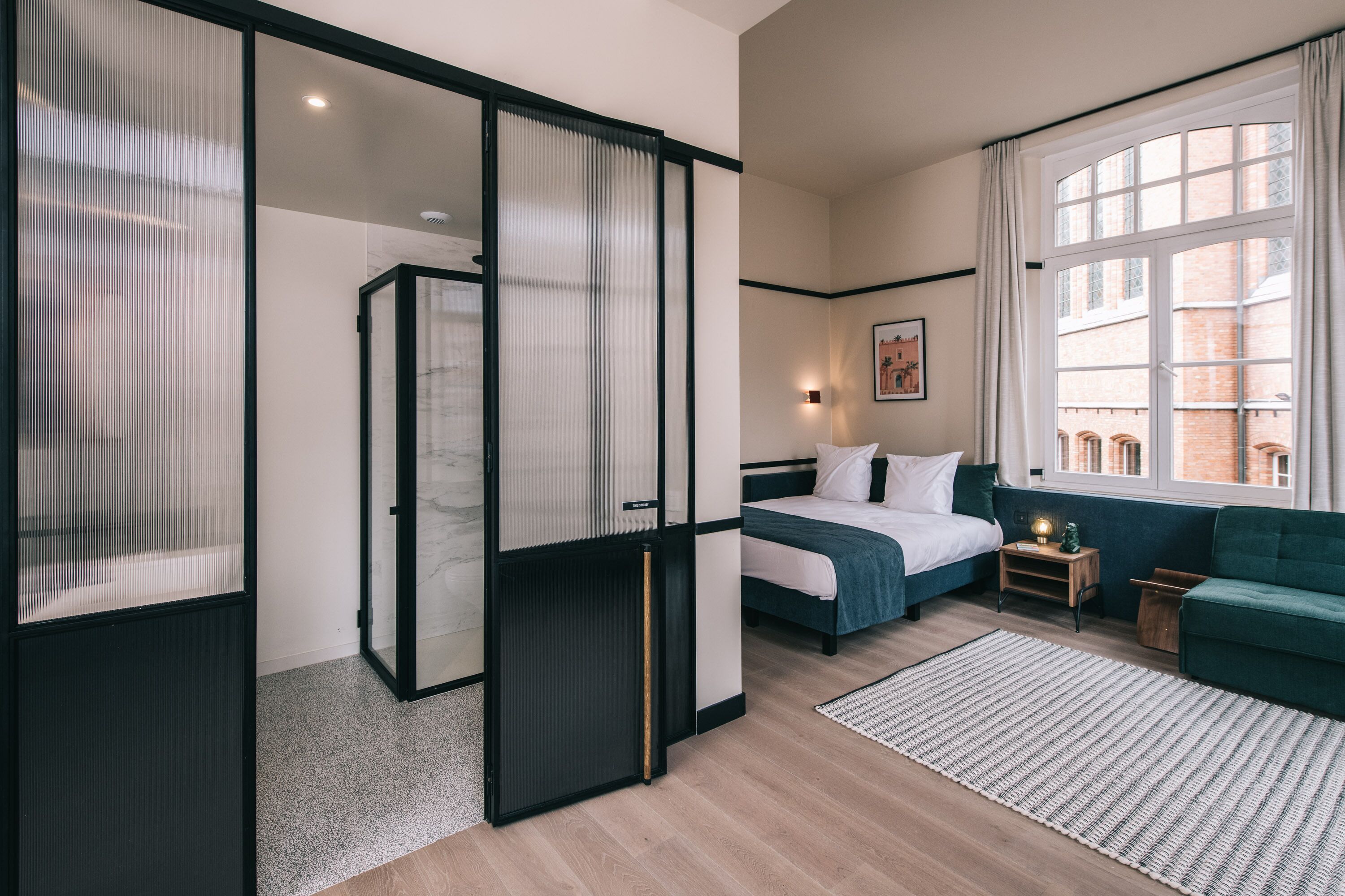 A modern hotel room with a bed, a sofa, a small table with a lamp, and a large window with curtains.