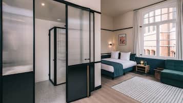 A modern hotel room with a bed, a sofa, a small table with a lamp, and a large window with curtains.