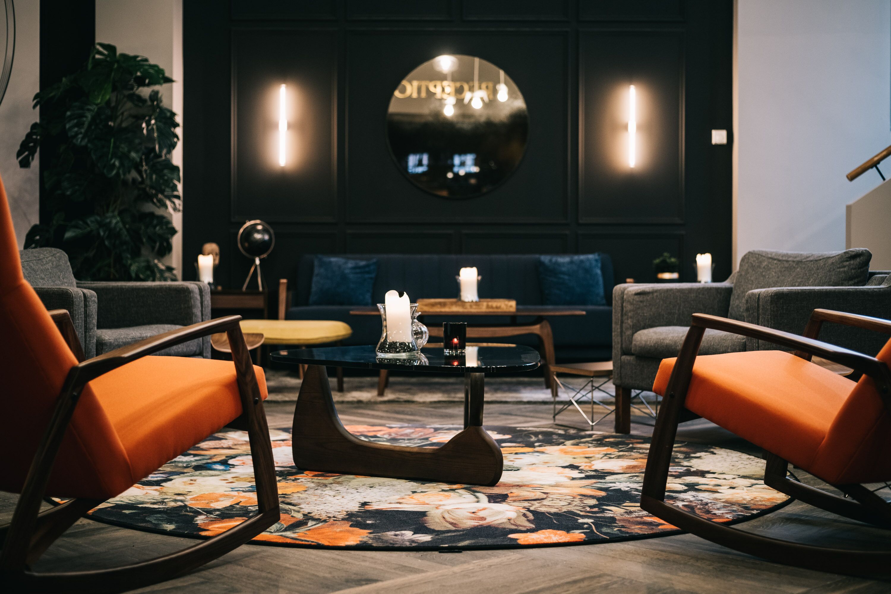 Een moderne zithoek met een ronde tafel, twee oranje fauteuils, een bank en een grote spiegel aan de muur.