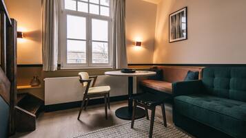 A cozy room with a green sofa, a round table, and a chair. There is a window with curtains, a radiator, and a wall-mounted light.