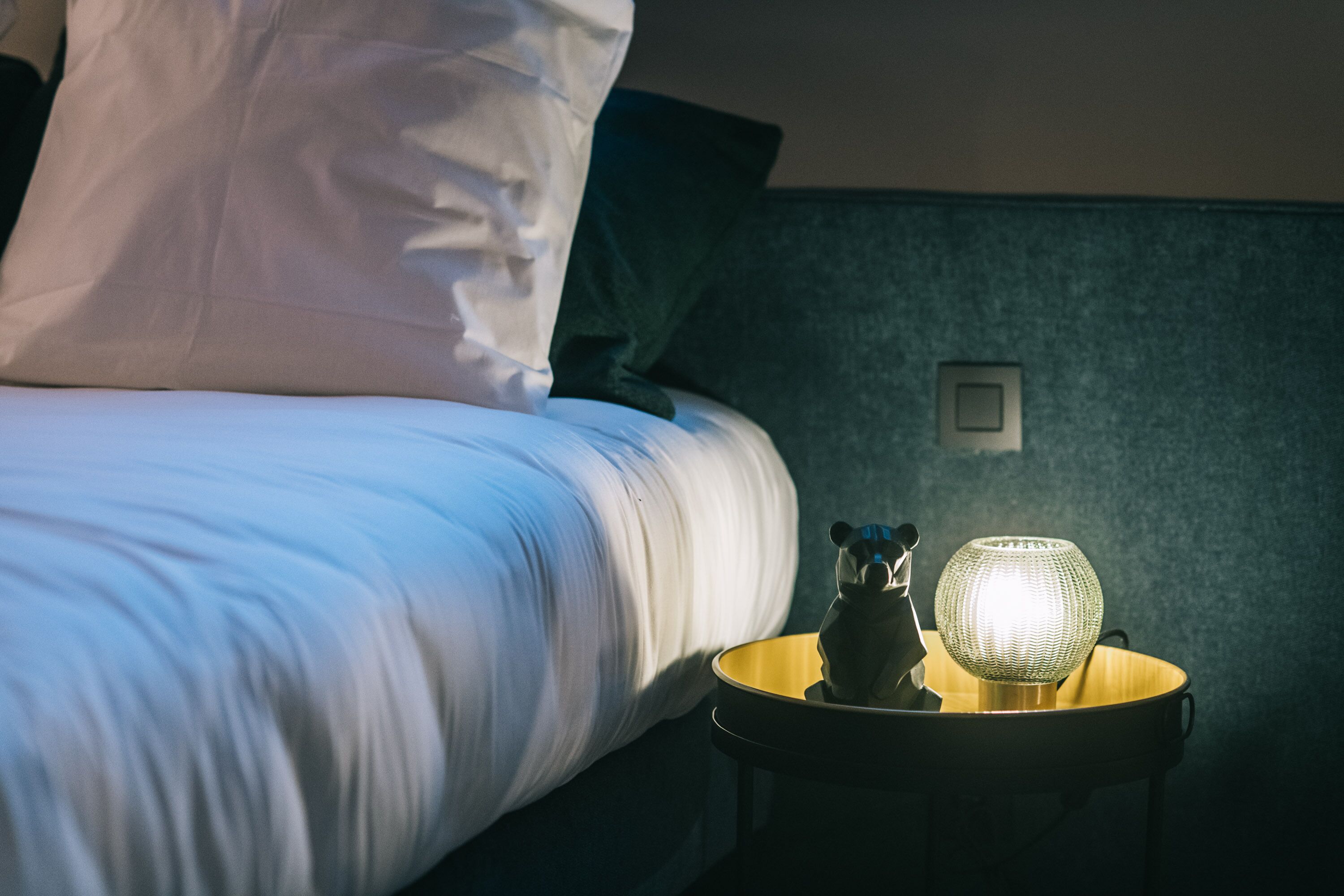 A neatly made bed with white linens, a small table with a lamp, and a wall socket.
