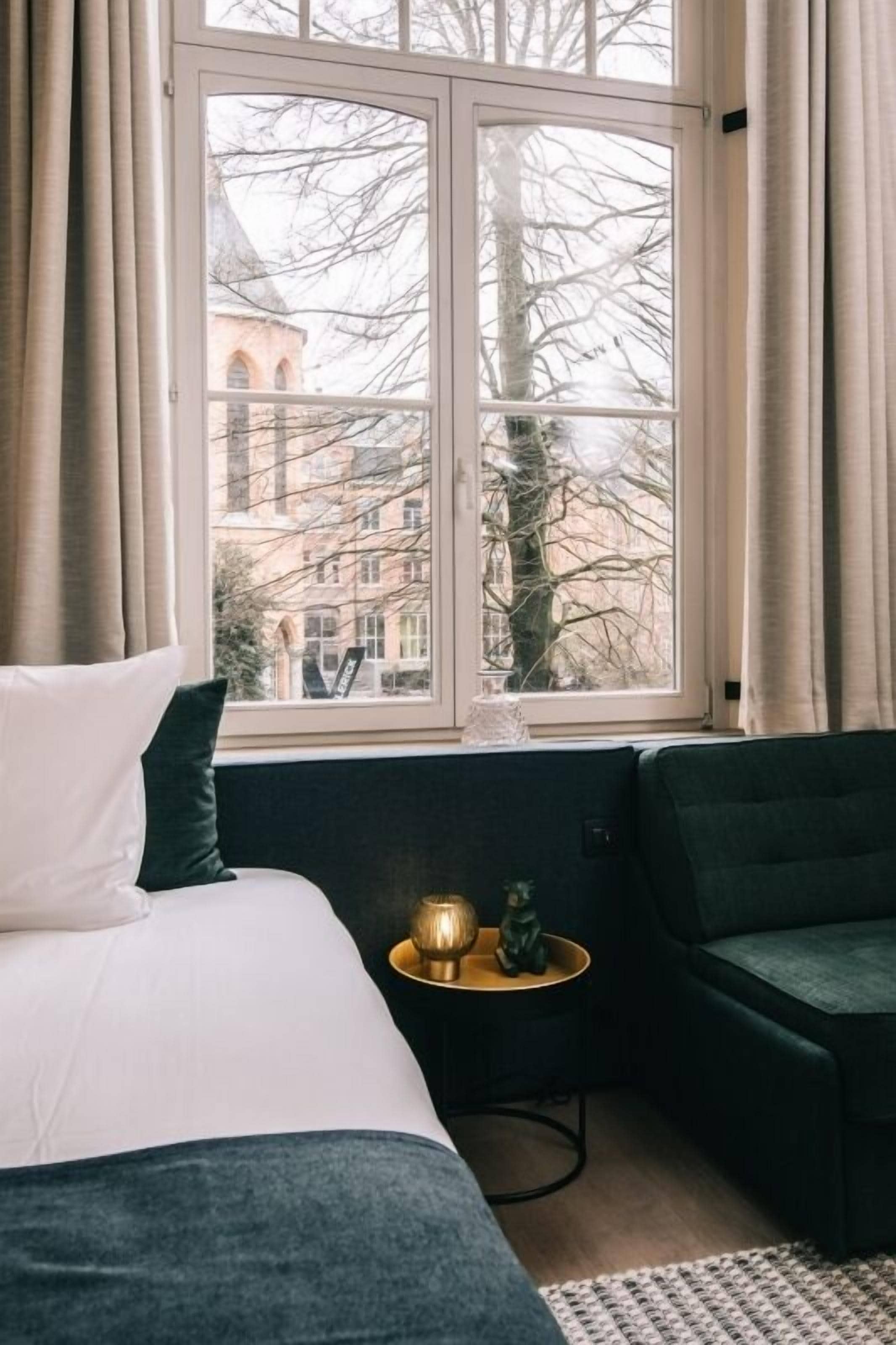 Un dormitorio con un ventanal grande, una cama con ropa de cama blanca y verde oscuro, una mesita redonda con un objeto decorativo y un sillón verde oscuro.