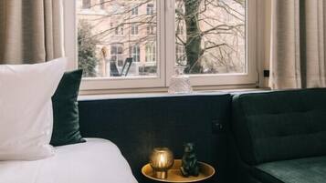 Een slaapkamer met een groot raam, een bed met wit en donkergroen beddengoed, een klein rond tafeltje met decoratie, en een donkergroene fauteuil.
