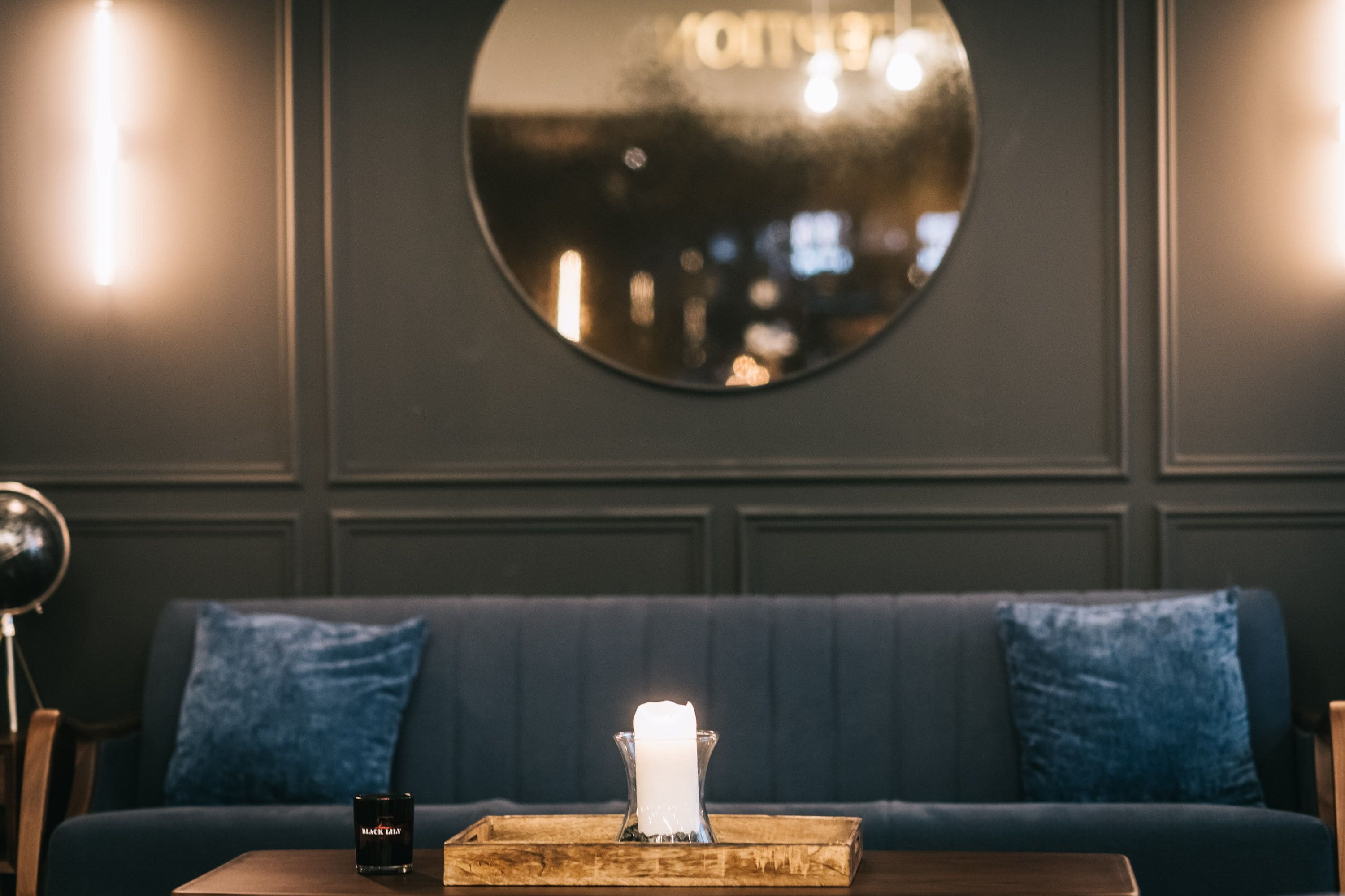 Une pièce aux couleurs sombres, avec un grand miroir rond accroché au mur, un canapé bleu agrémenté de coussins assortis, une table en bois sur laquelle se trouvent une bougie et un récipient noir.