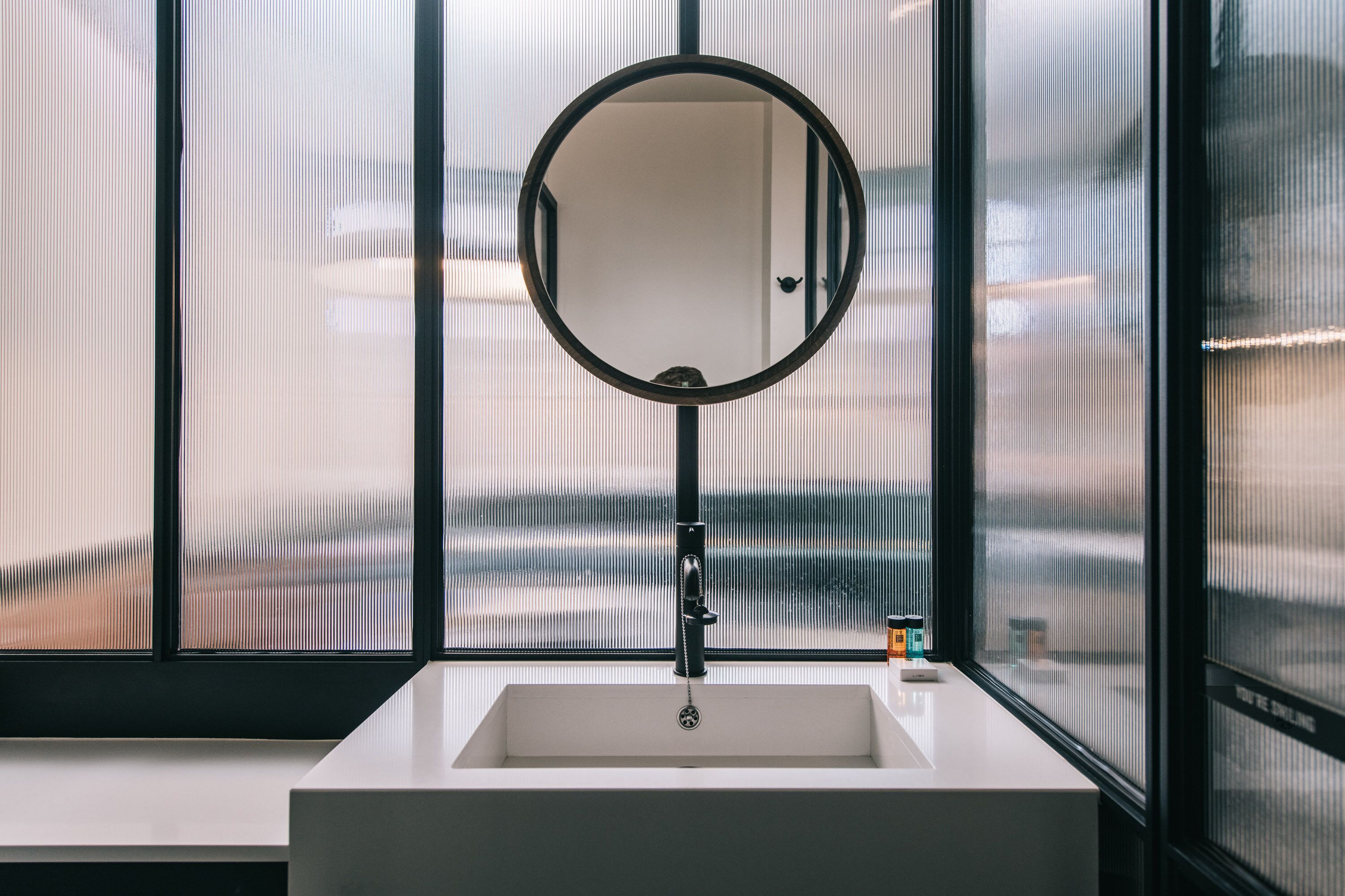 Un baño moderno con un ventanal amplio, un espejo redondo y un lavabo blanco.