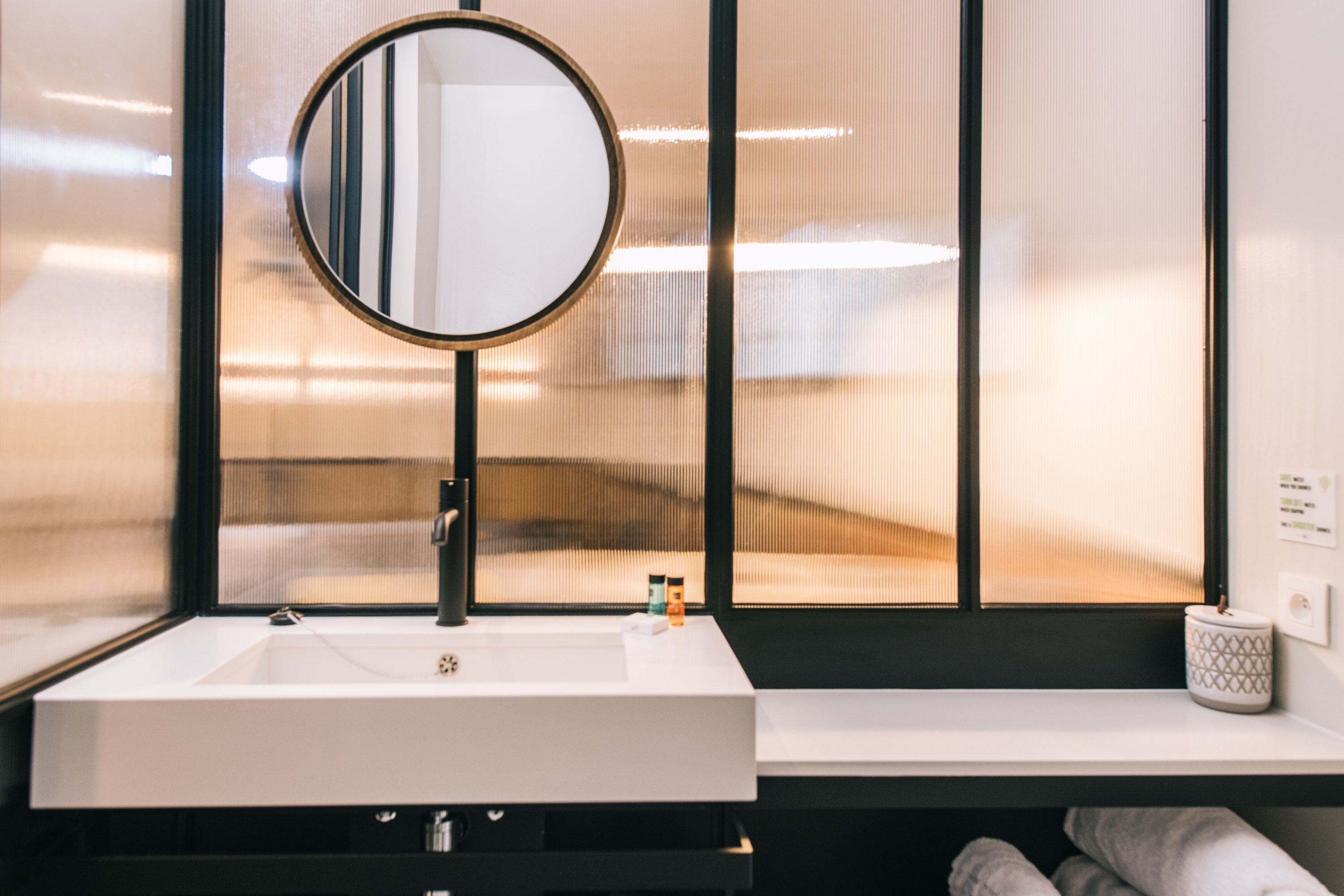Een moderne badkamer met een witte wasbak, een ronde spiegel en een raam met zonwering.