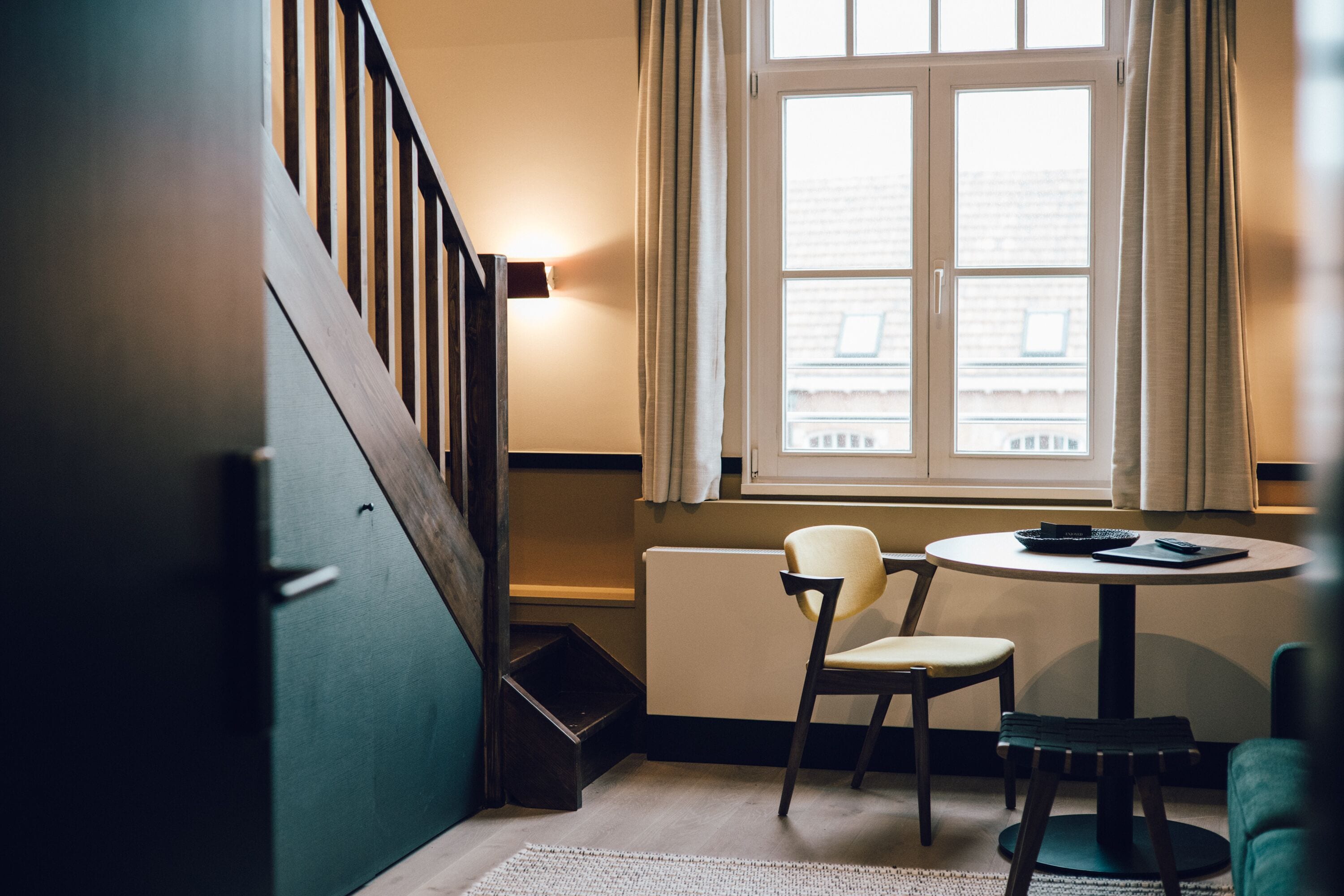 Une pièce avec un escalier en bois, une table ronde entourée de chaises, un radiateur et une fenêtre avec des rideaux.