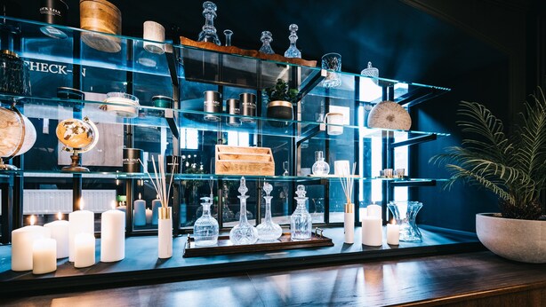 A bar area with shelves displaying various bottles and glasses, a potted plant, and candles.