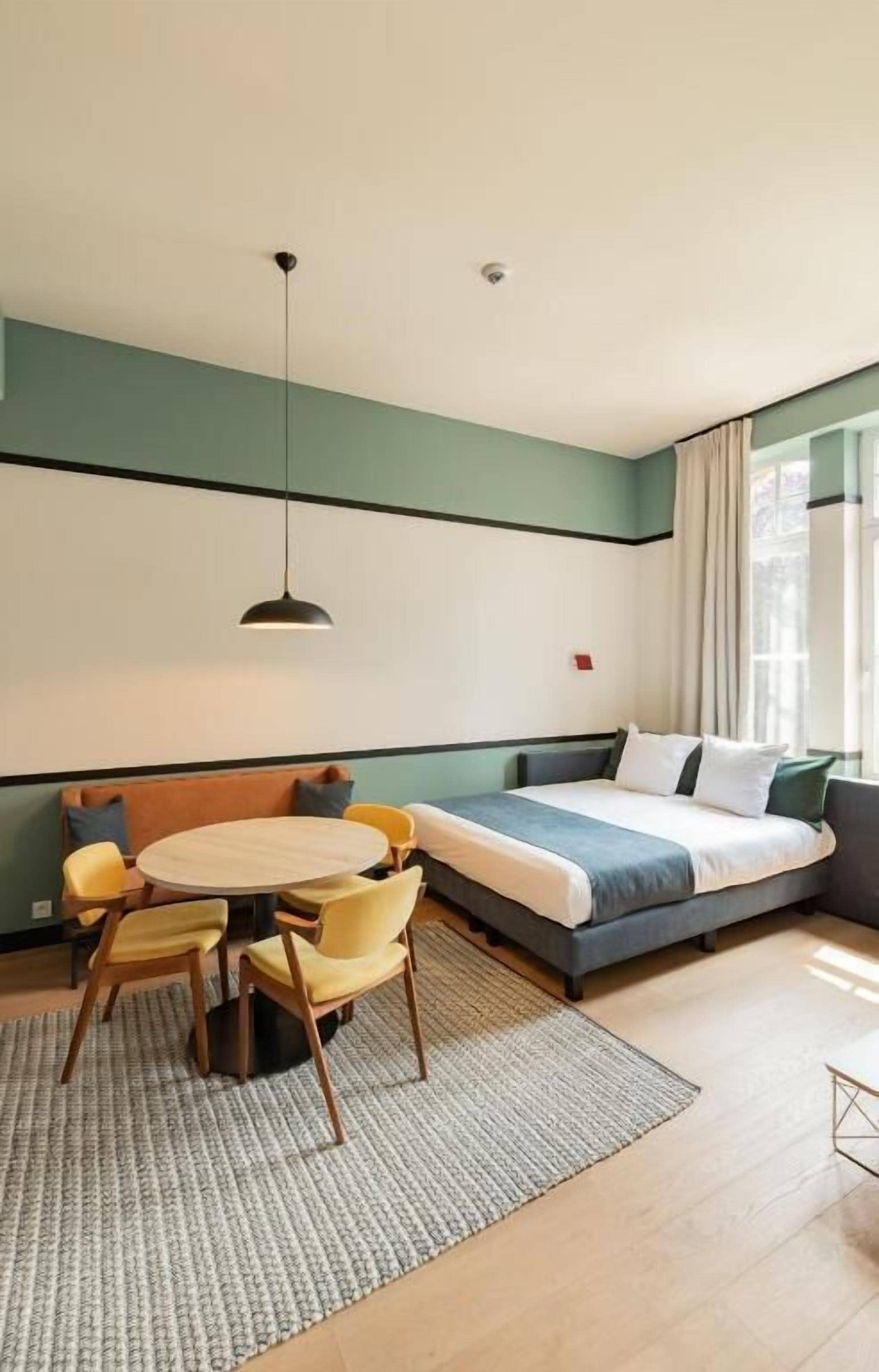 A modern hotel room with a bed, a sofa, a dining table with chairs, and a hanging light fixture.