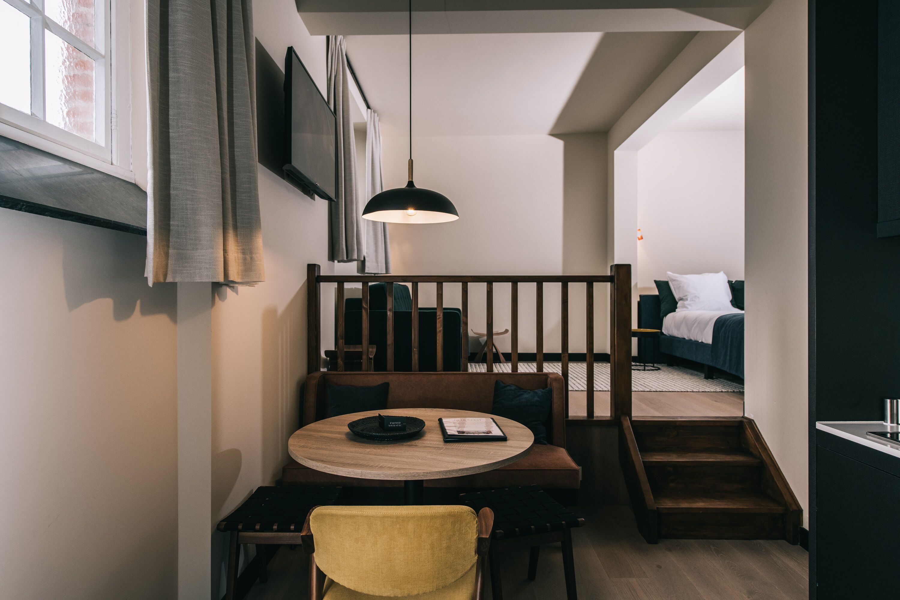 Ein modernes Interieur mit einem runden Holztisch, einem gelben Stuhl und einer schwarzen Pendelleuchte. Eine Treppe führt zu einem Schlafzimmer mit einem Bett und einem Nachttisch.
