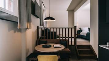 Ein modernes Interieur mit einem runden Holztisch, einem gelben Stuhl und einer schwarzen Pendelleuchte. Eine Treppe führt zu einem Schlafzimmer mit einem Bett und einem Nachttisch.
