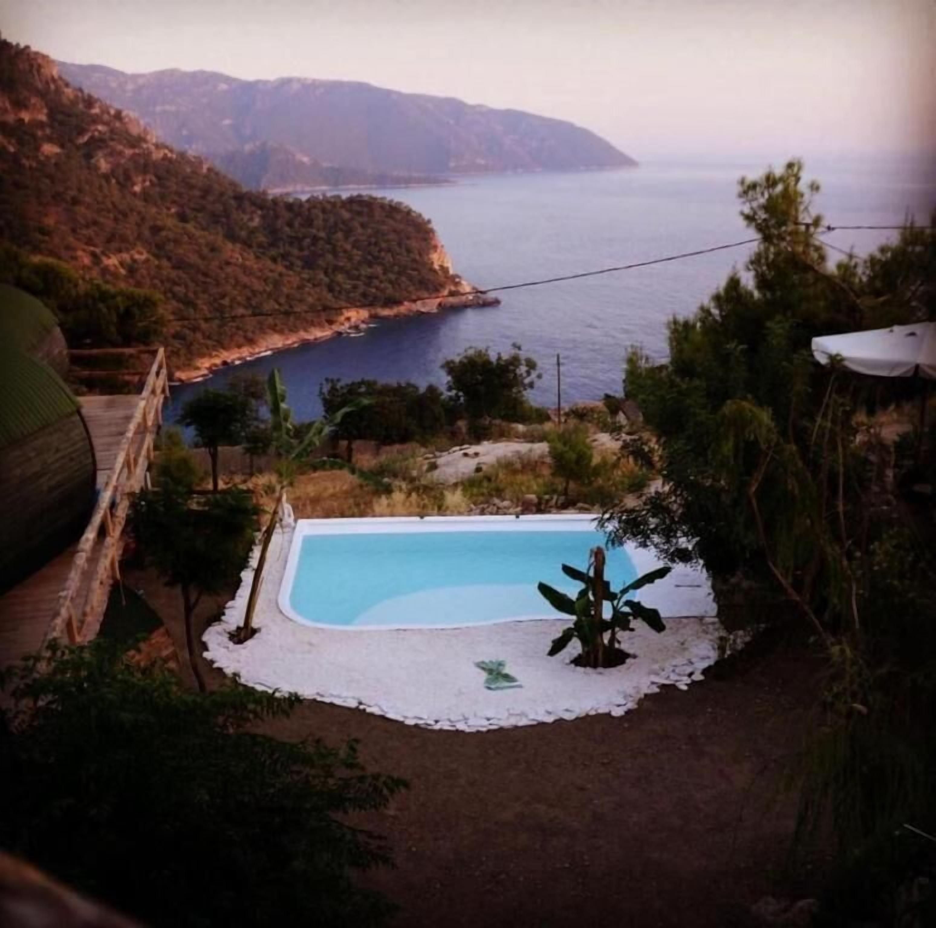 A swimming pool surrounded by a white deck, with a view of the sea and hills in the background.
