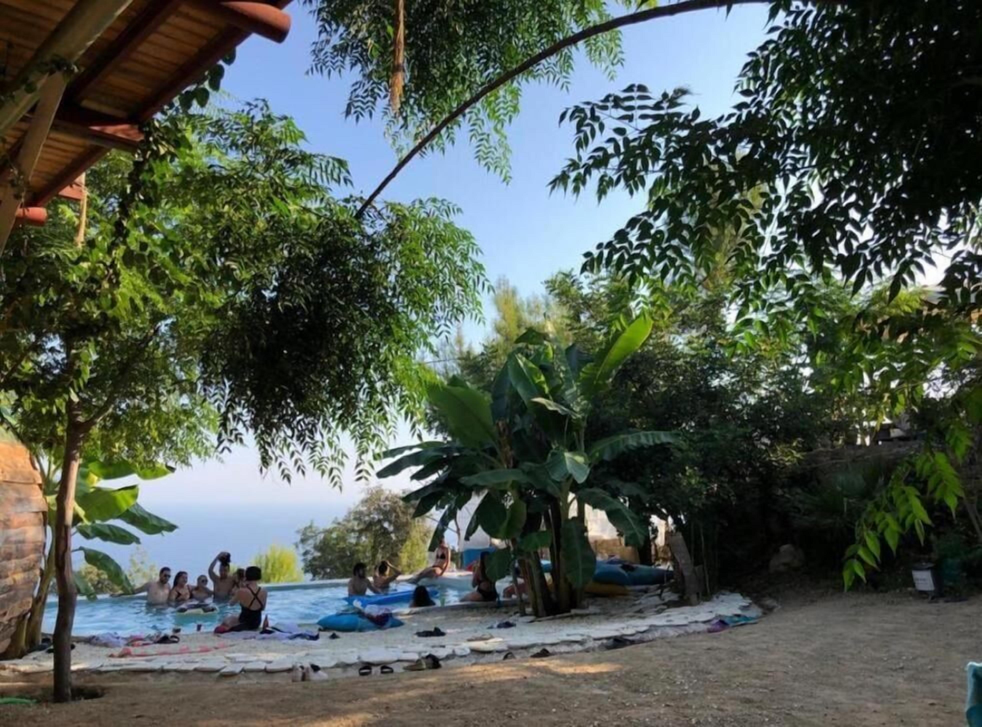 An outdoor swimming pool area with people relaxing and sunbathing, surrounded by trees and a building structure.