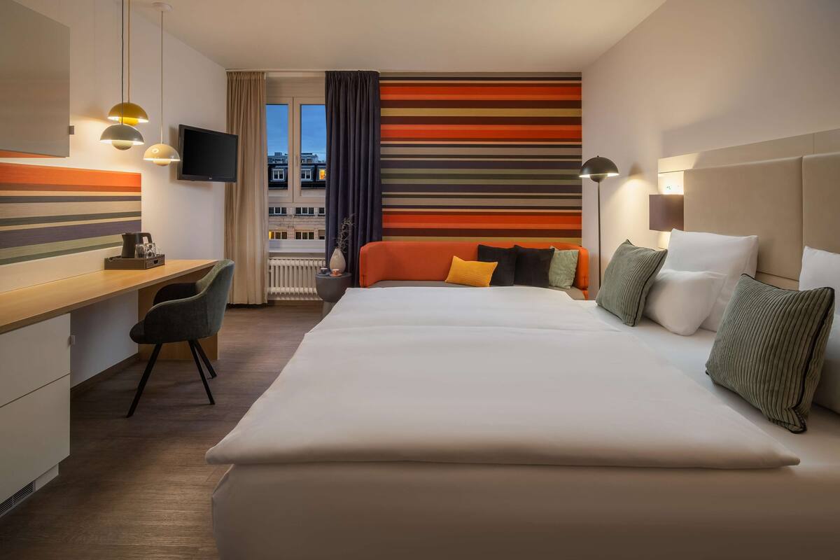 A hotel room with a large bed, a desk with a chair, a TV, and a view of buildings outside.