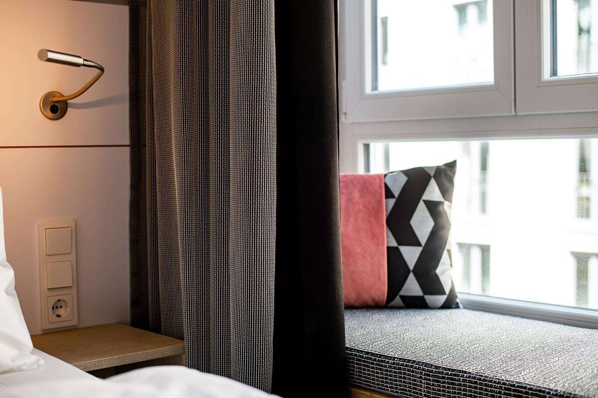 A hotel room with a bed, a window with a view, a curtain, and a wall-mounted light fixture.