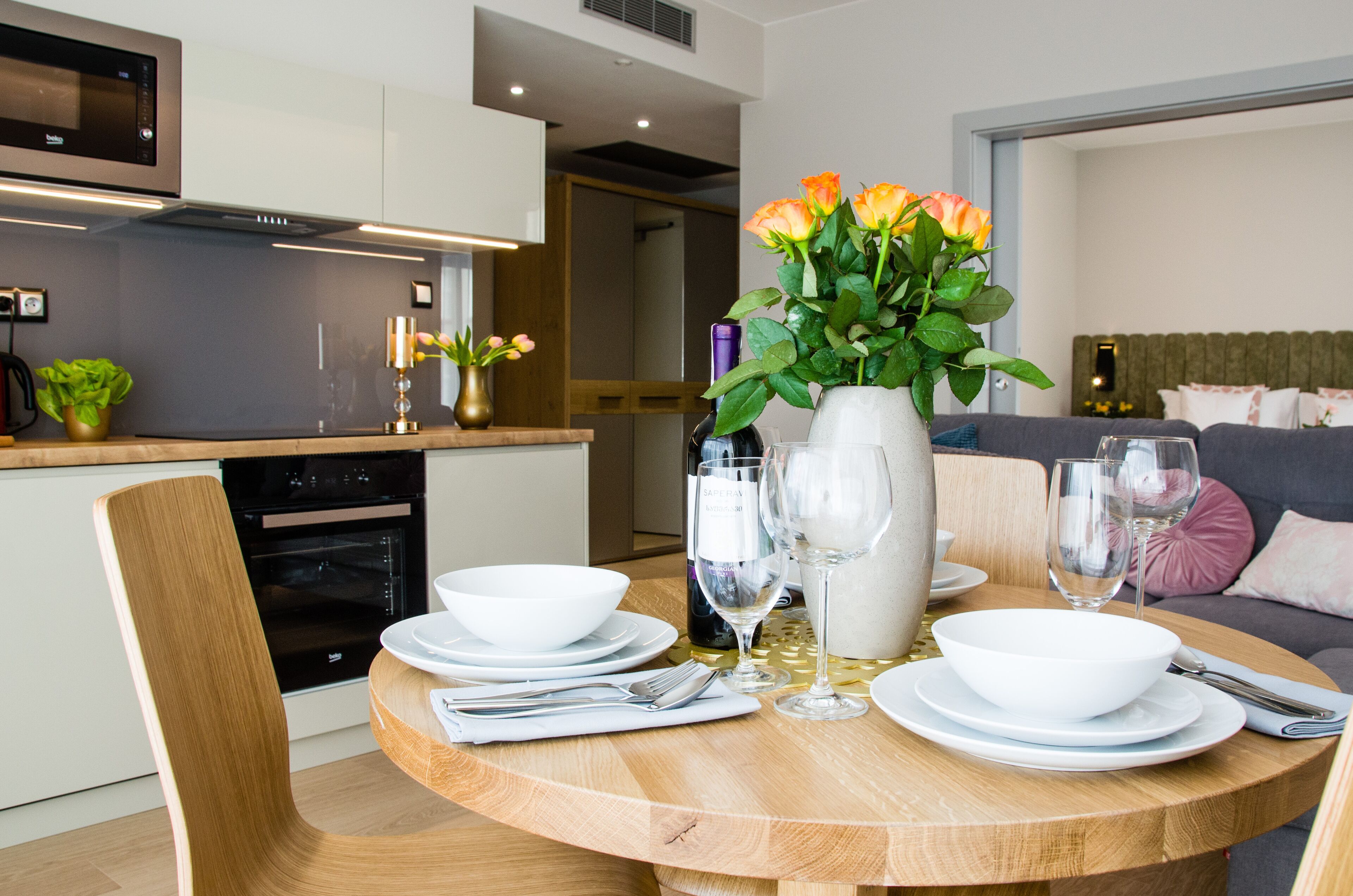 Een moderne eethoek met een ronde houten tafel gedekt voor twee personen, witte borden, bestek en een vaas met bloemen.