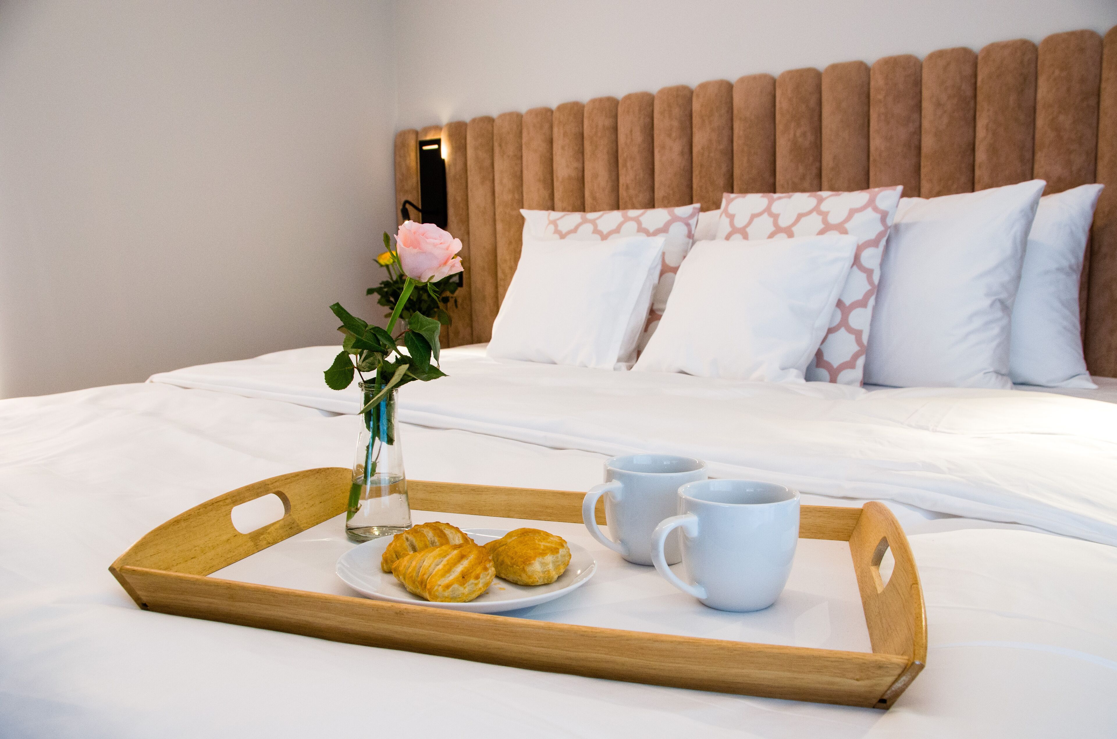 A neatly made bed with white linens, a wooden headboard, and decorative pillows. A tray with croissants and coffee cups is placed on the bed.