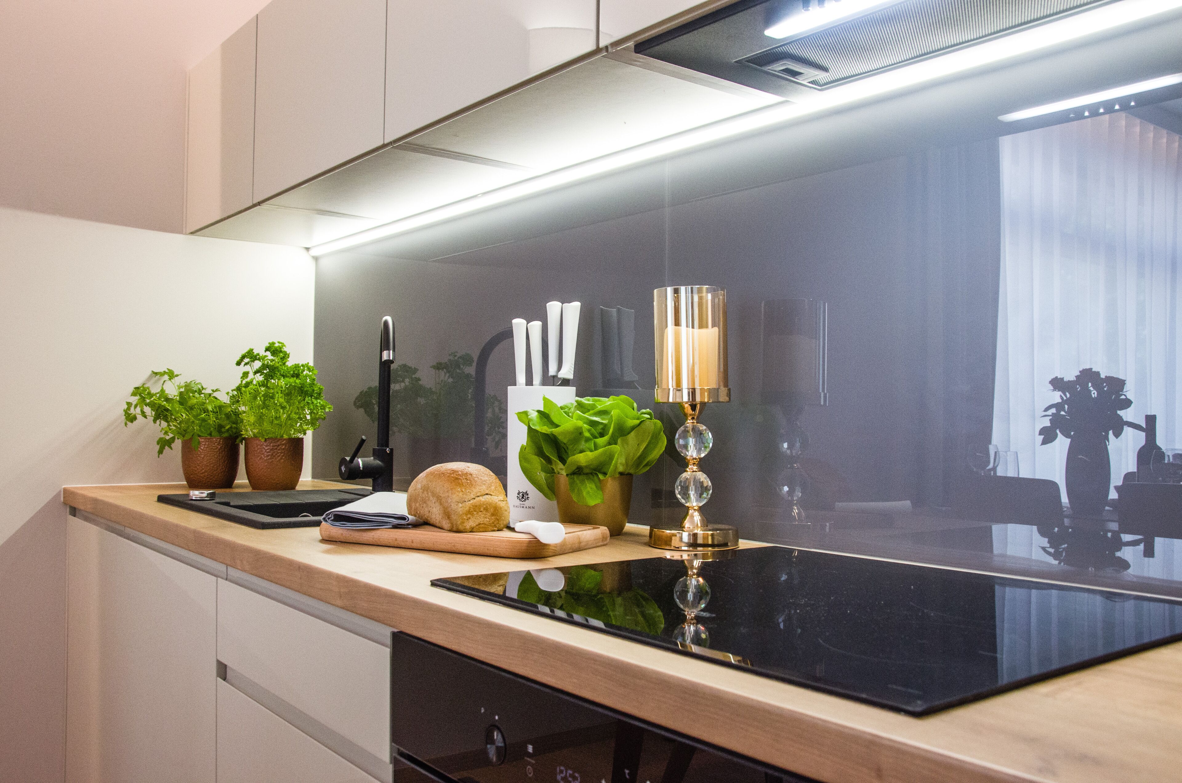 A modern kitchen with a black induction cooktop, wooden countertop, potted plants, and a decorative candle holder.
