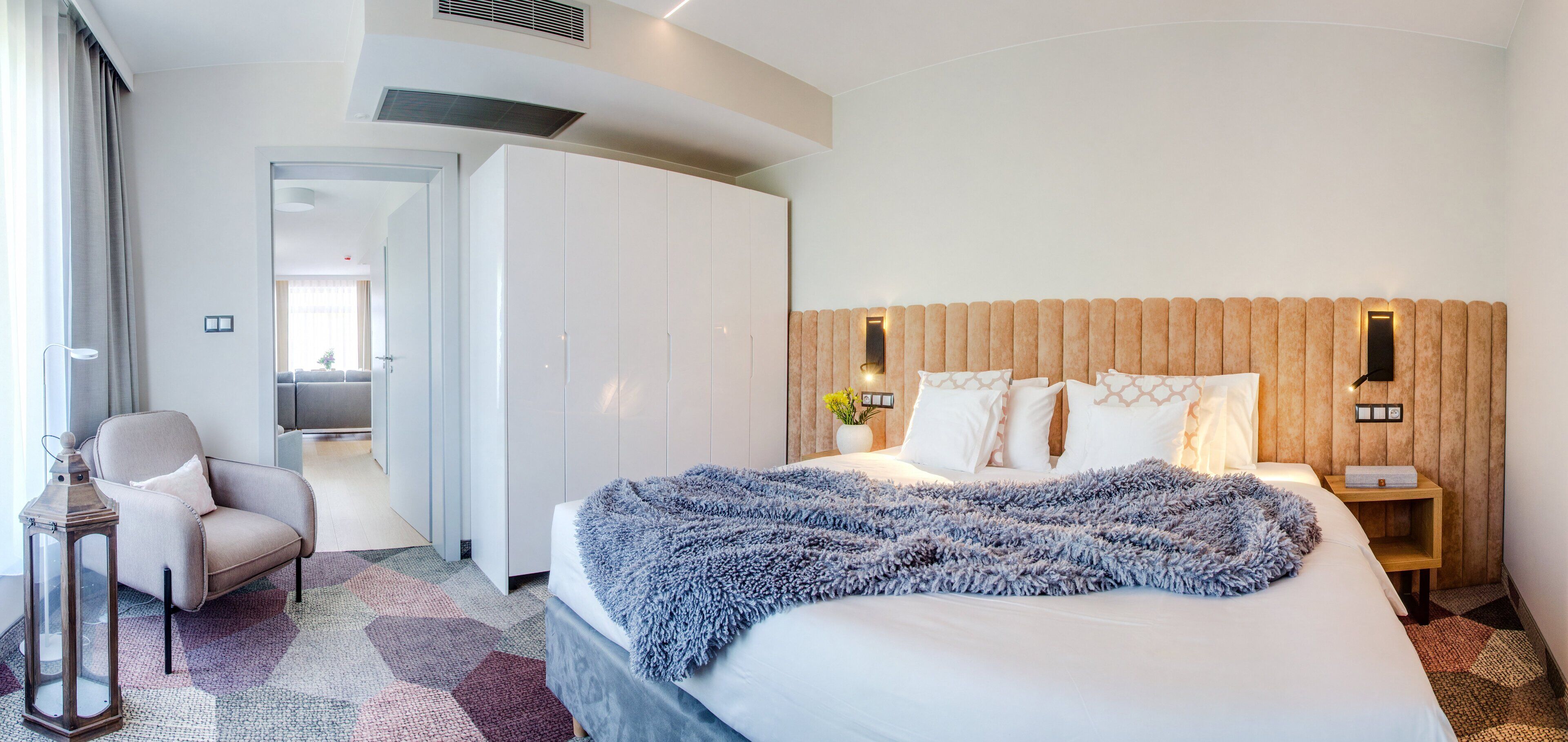 A hotel room with a large bed, a chair, a wardrobe, and a bedside table with a lamp.