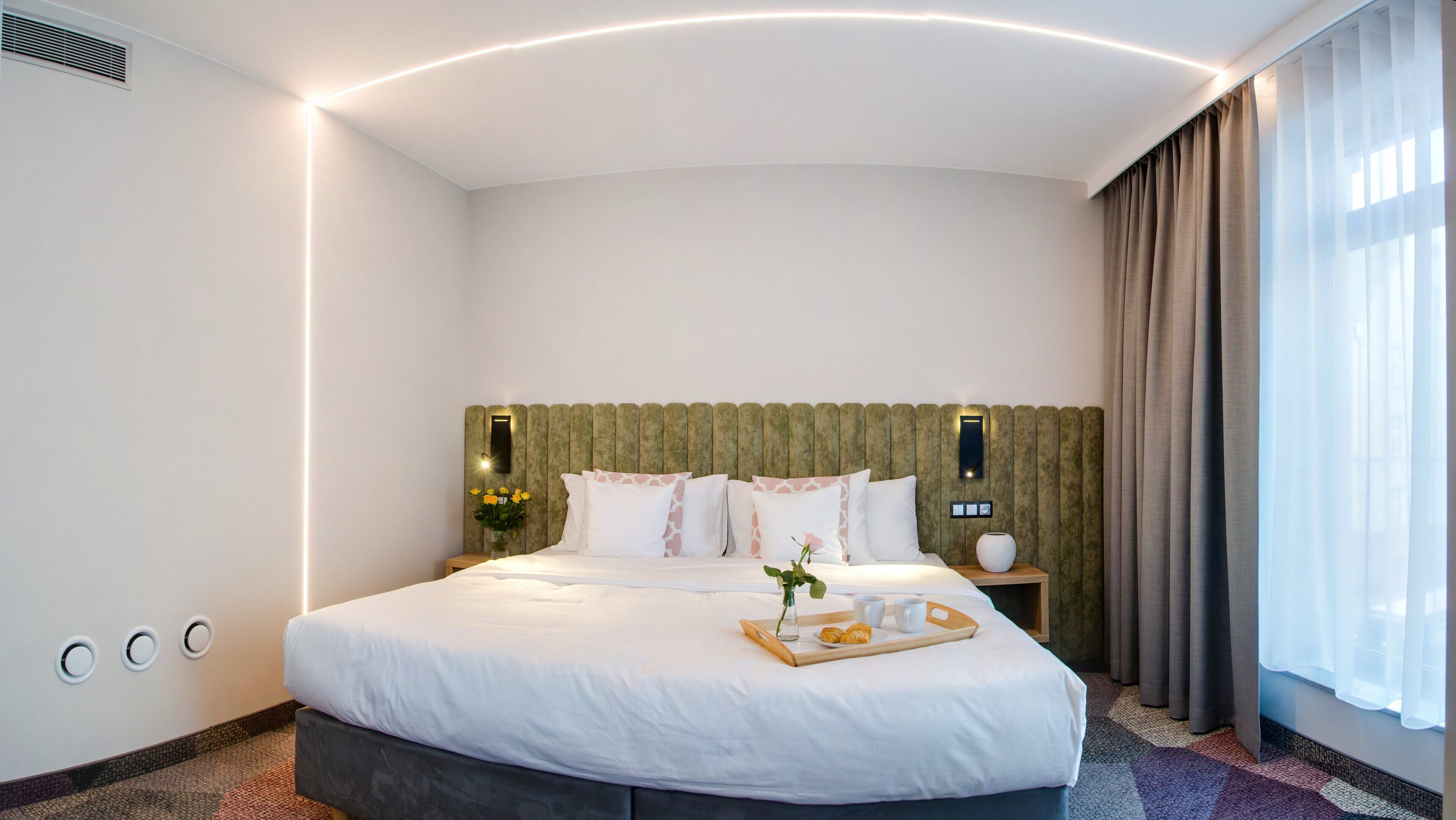 A modern hotel room with a large bed, a bedside table with a vase and a tray of pastries, and a view of the city through sheer curtains.