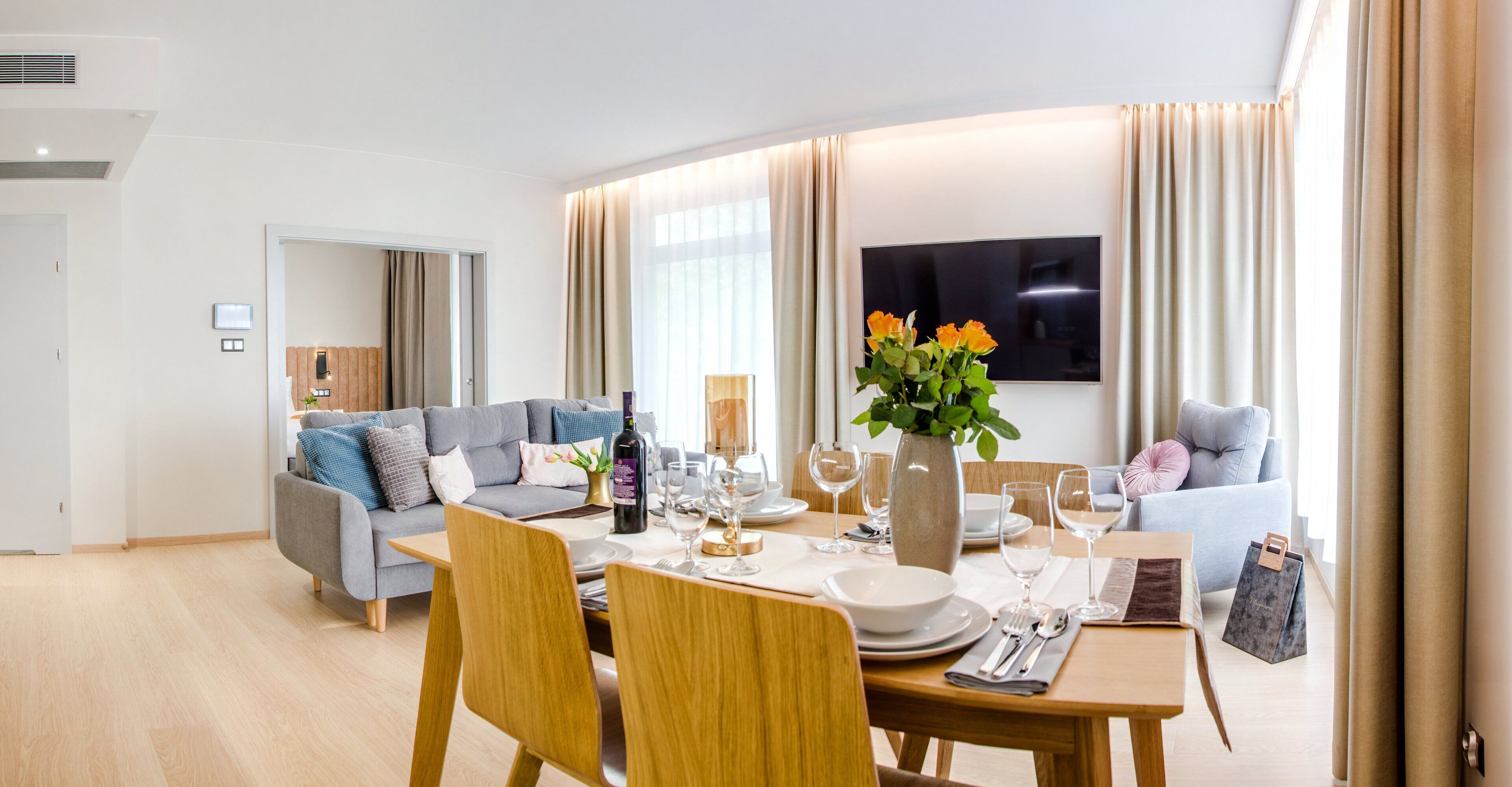 A modern living room with a dining area, a grey sofa, a wooden dining table, and a flat-screen TV mounted on the wall.