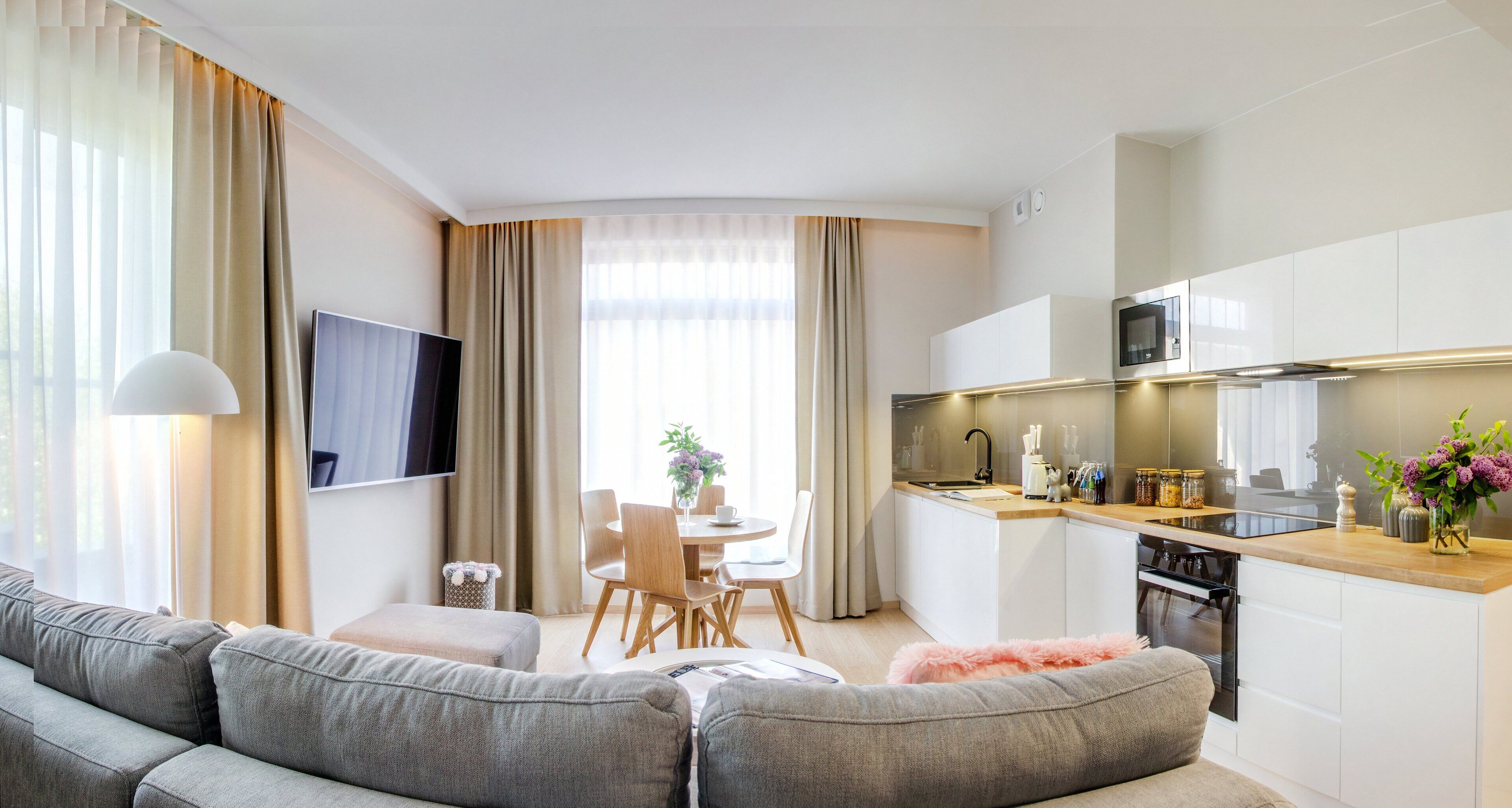 A modern living room with a grey sofa, a dining area with a table and chairs, and a kitchen with white cabinets and a microwave.